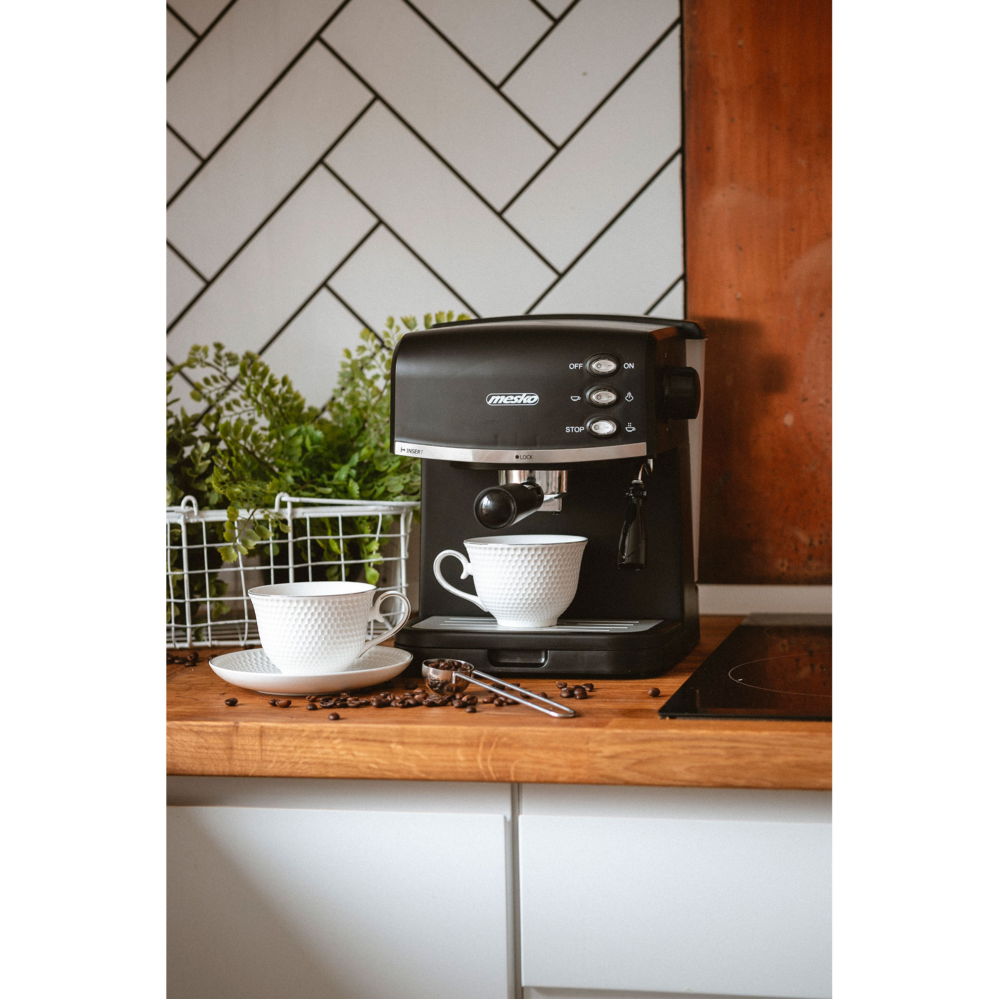 Ekspres do kawy z dwoma białymi filiżankami i łyżeczką, z ziarnami kawy w tle.