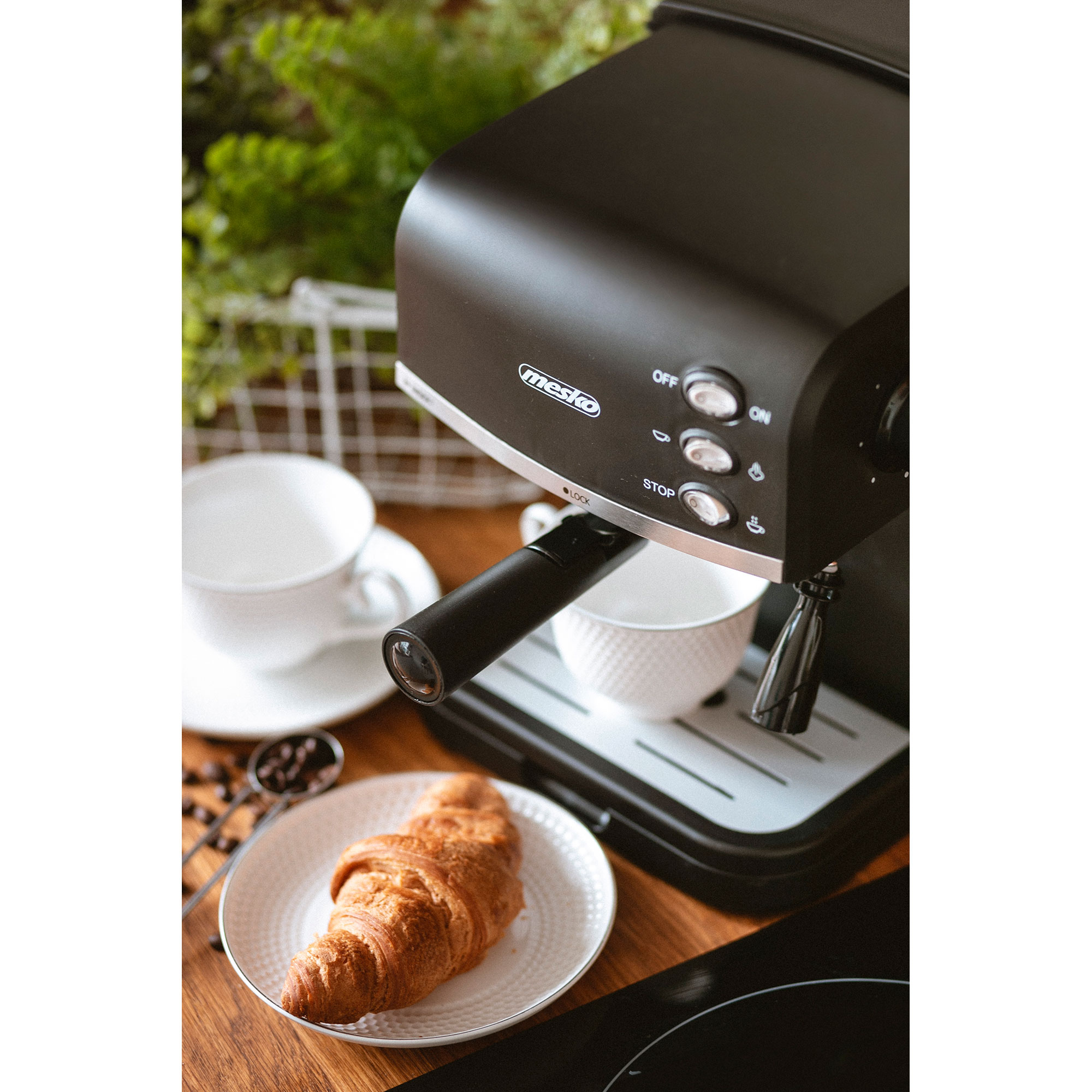 Ekspres do kawy z filiżankami, rogalikiem i ziarnami kawy na drewnianej powierzchni.