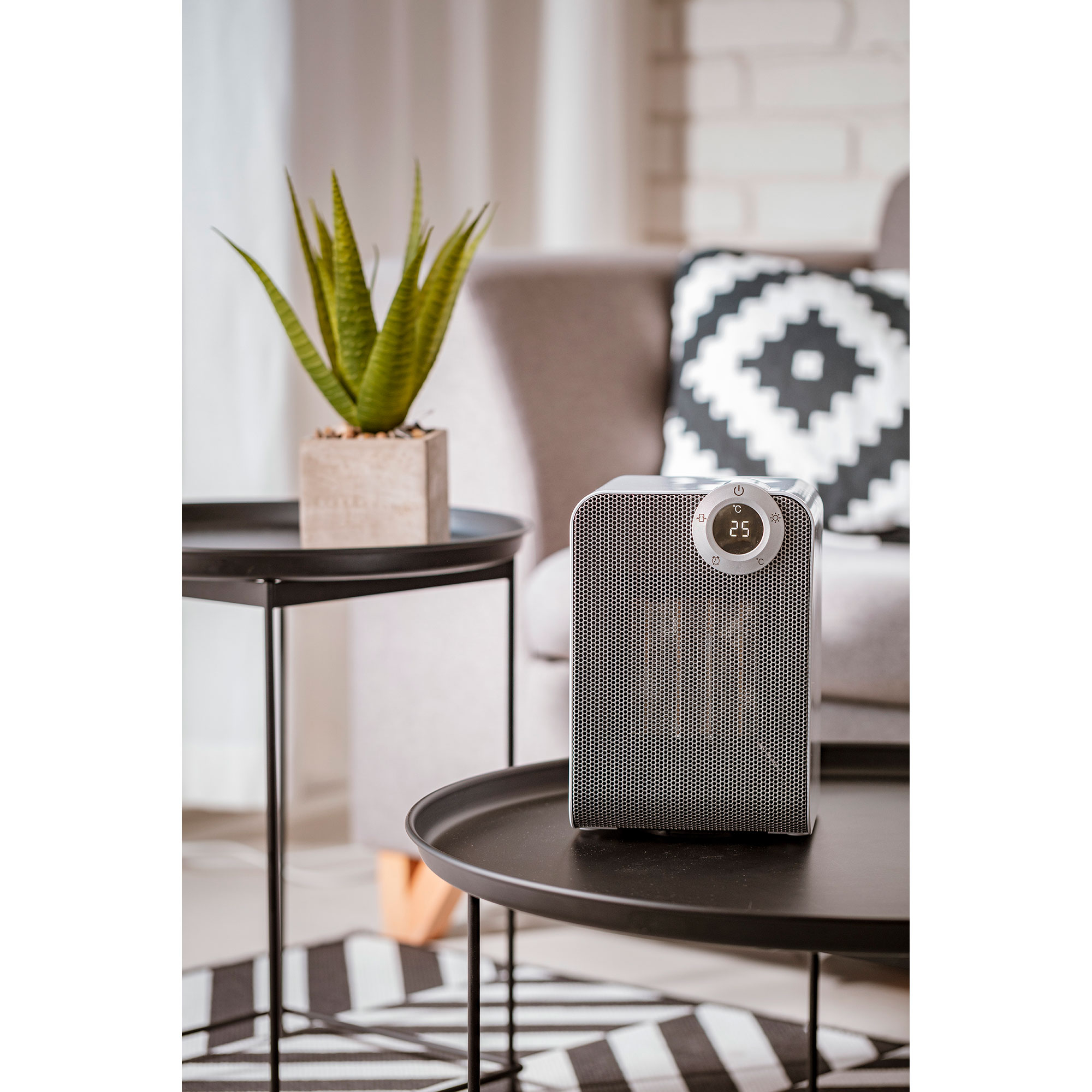 Kleiner weißer Heizlüfter auf einem Tisch mit Digitalanzeige, anderer Tisch mit Pflanze und Sofa.