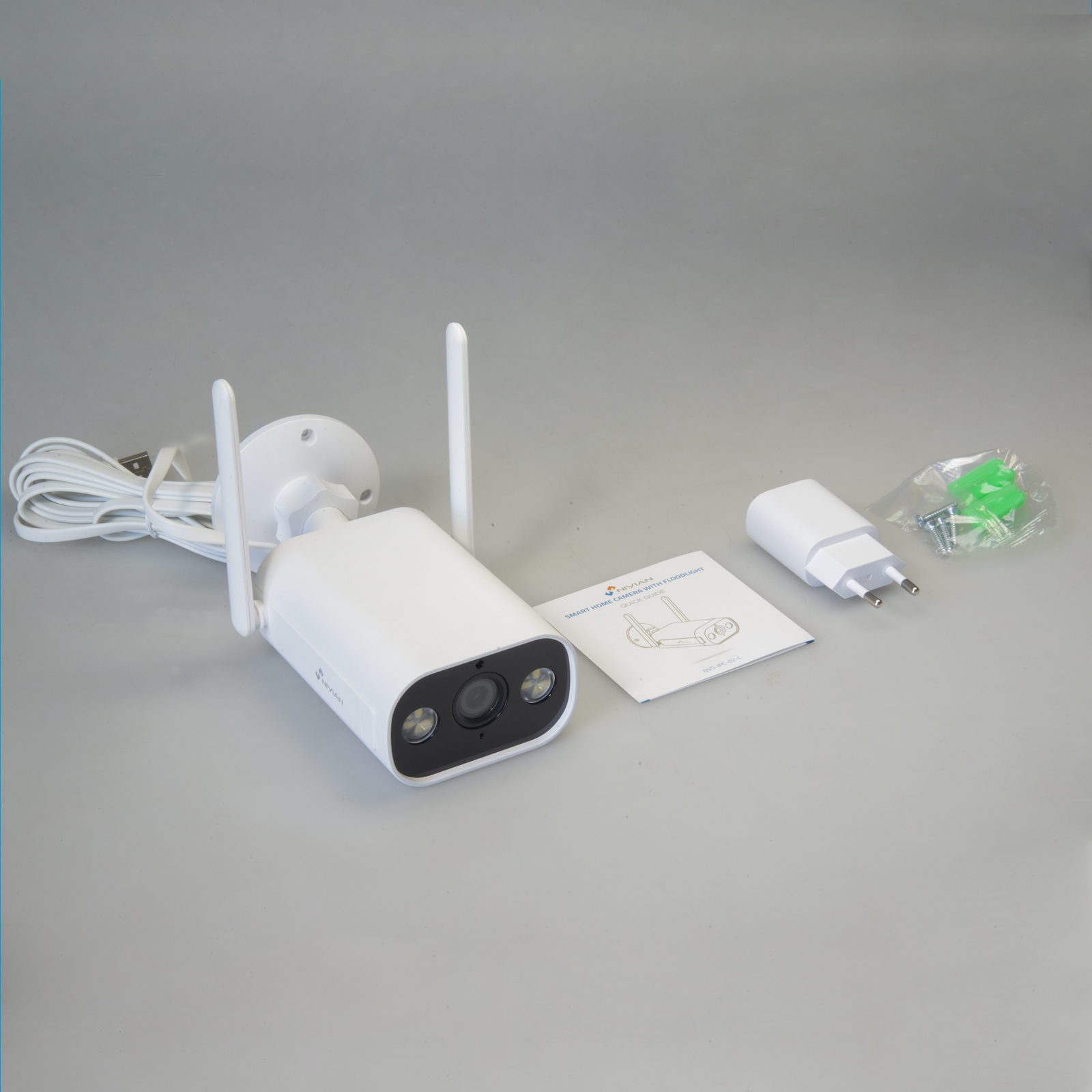 Una cámara de seguridad blanca con antenas, un cargador y un manual sobre una superficie gris.