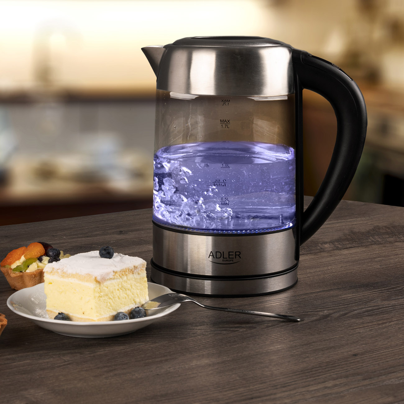 Szklany czajnik z wodą w środku, kawałek ciasta i talerz na drewnianym stole.