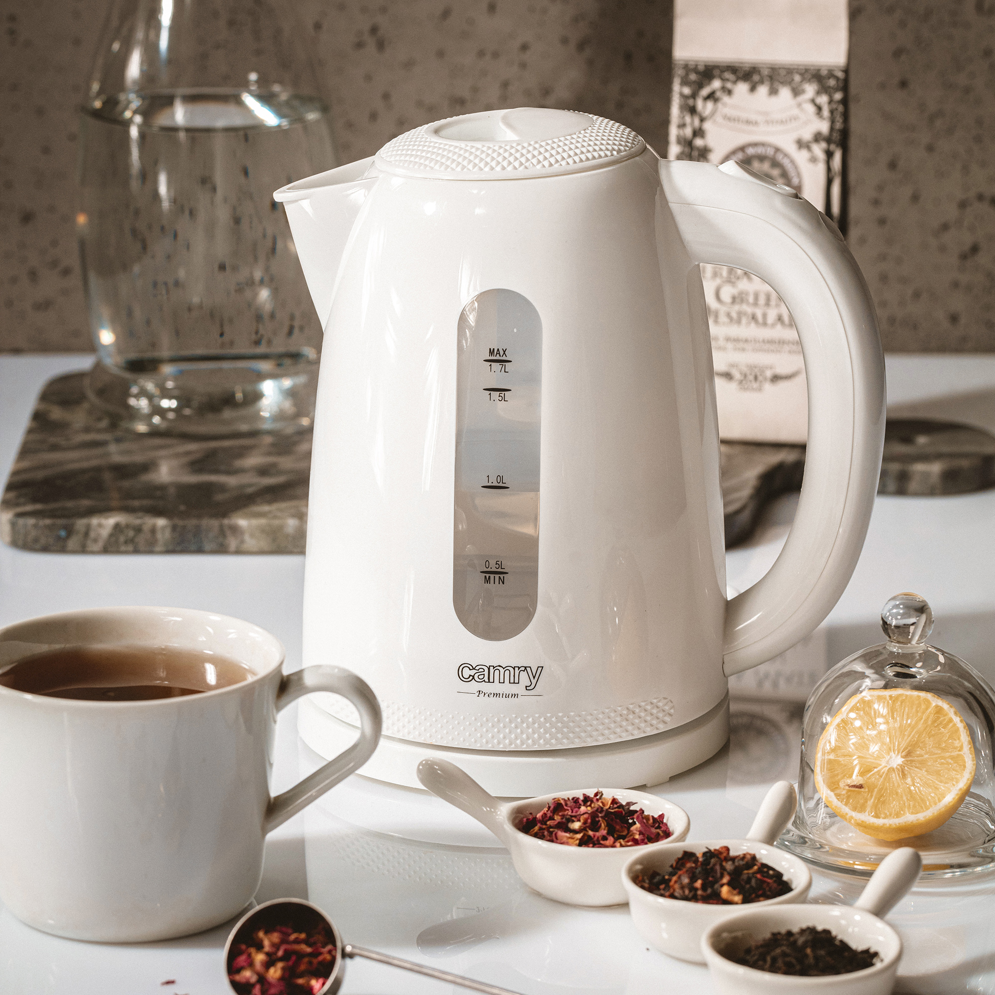 Hervidor eléctrico blanco sobre una mesa con té, limón y hierbas. El fondo tiene agua en un vaso.