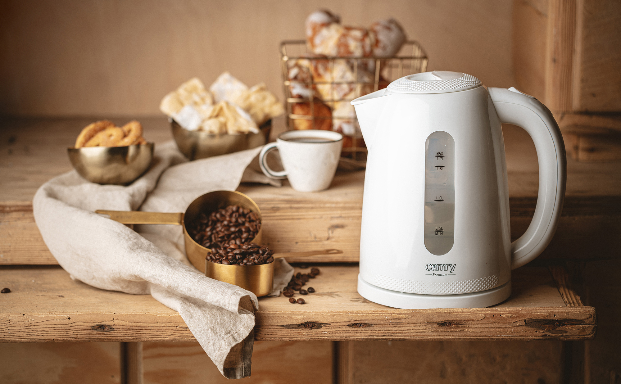 Un hervidor eléctrico blanco con ventana medidora. Granos de café, pasteles y una taza sobre una mesa de madera.