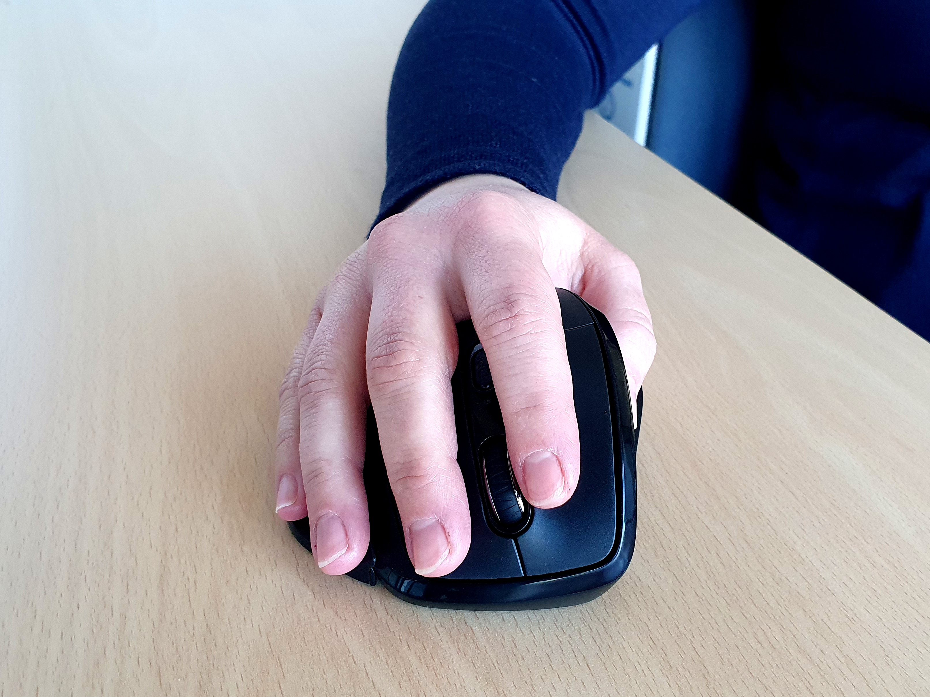 Eine Hand mit dunkelblauem Ärmel hält eine schwarze Computermaus auf einem hellen Holztisch.