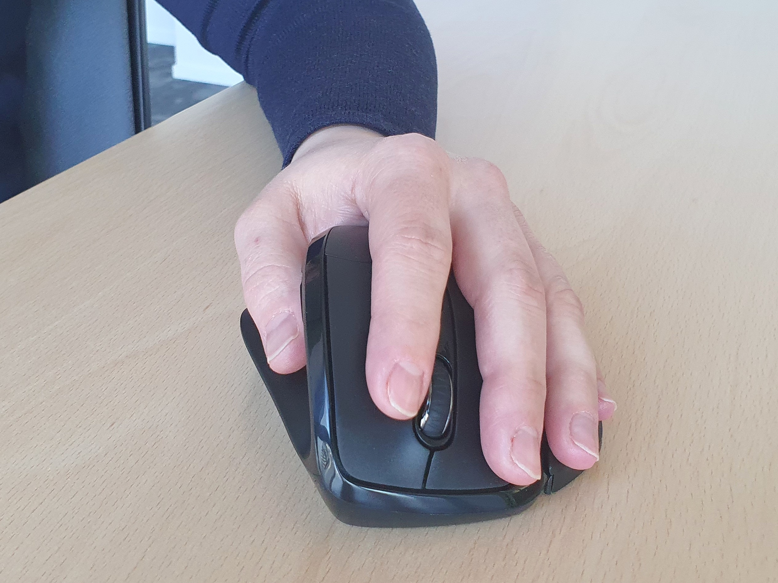 Die Hand einer Person, die eine schwarze, ergonomische Maus hält. Die Maus liegt auf einer hellen Holzoberfläche.