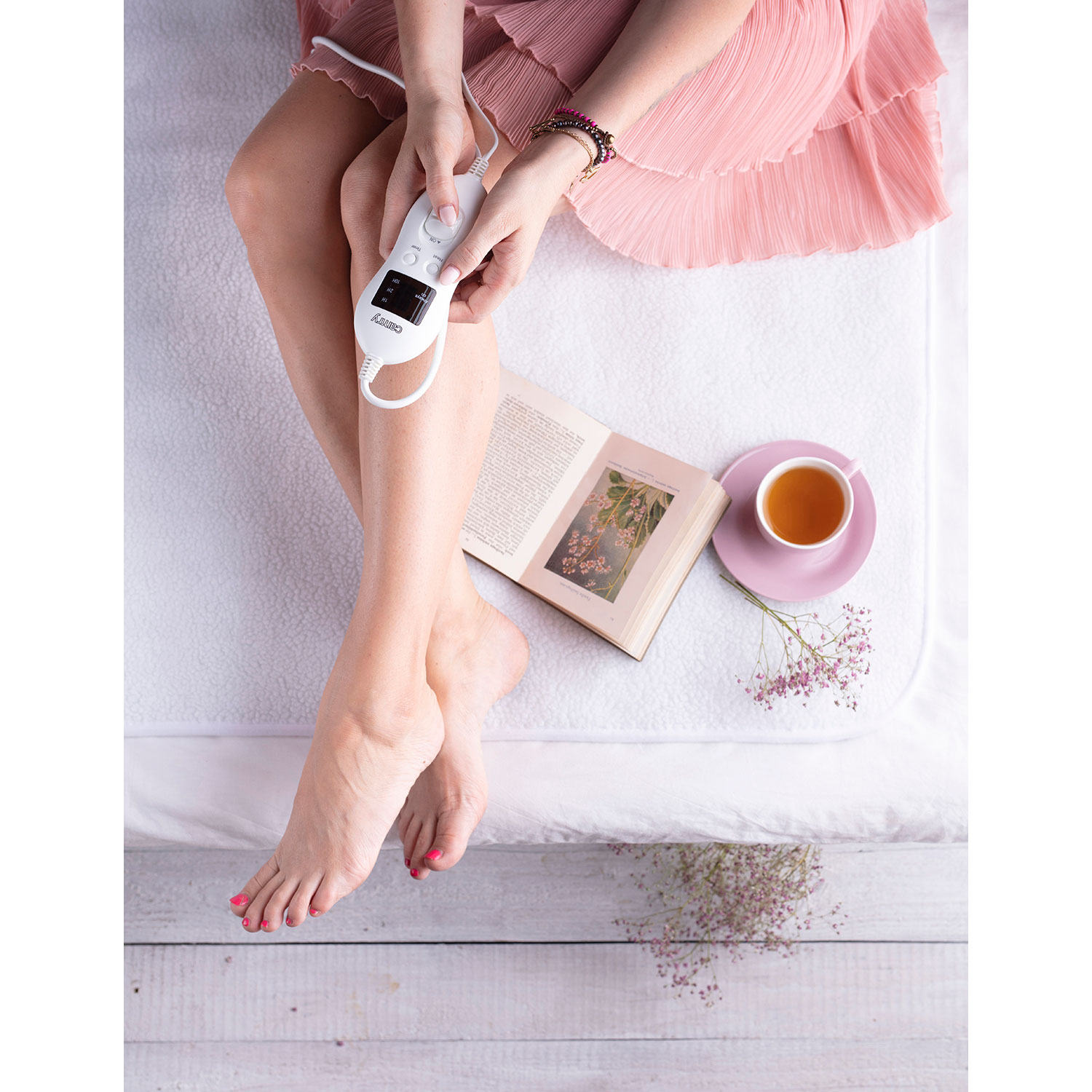 Eine Frau mit pinken Nägeln und einem rosa Kleid hält einen Controller, ein Buch und Tee auf einem Bett.