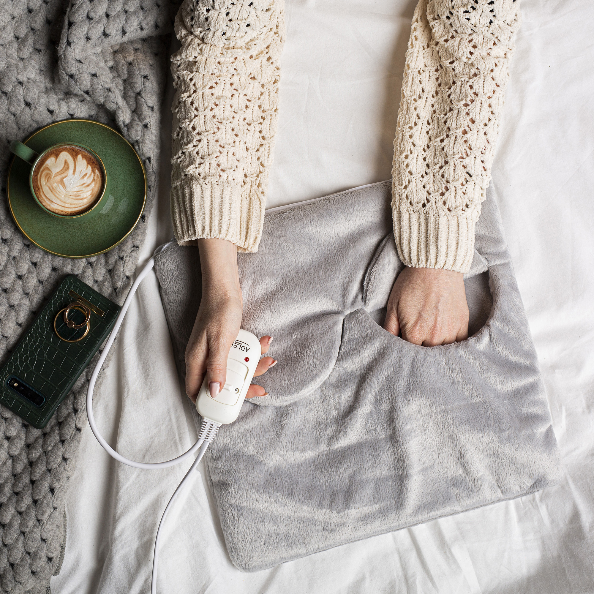 Una persona usando un calentador de pies gris en una cama. Hay un teléfono y una taza de café cerca.