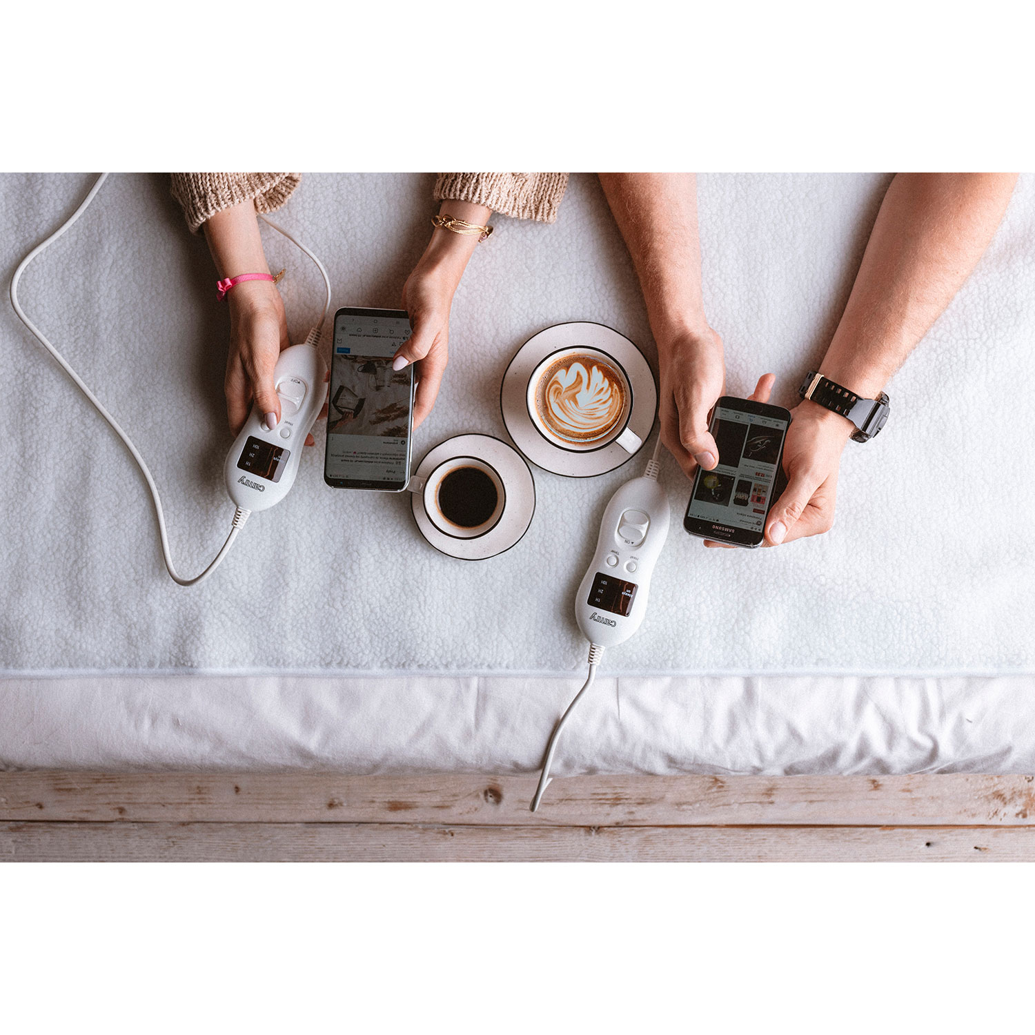 Two people on a bed with phones and coffee. They have heating pads with controllers.