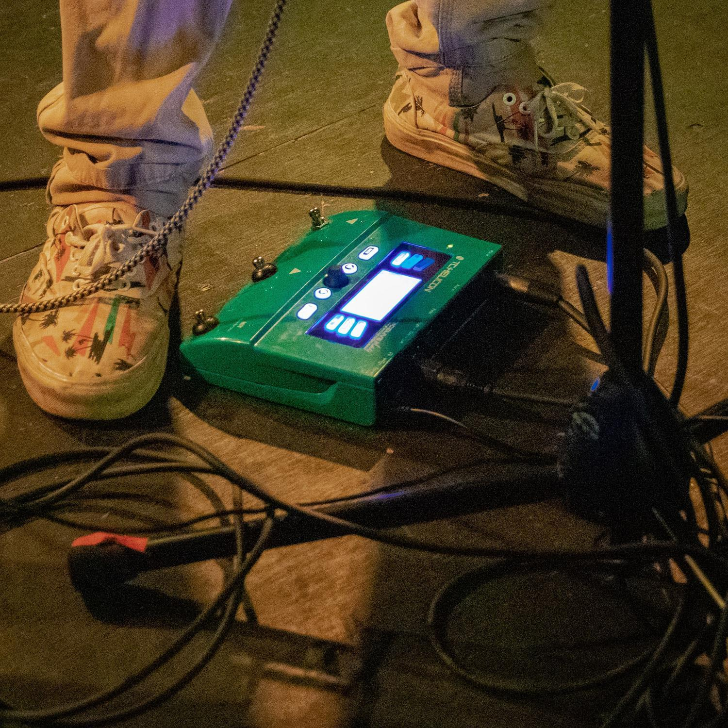 Personenfüße mit Schuhen auf einer Bühne, grünes Musikpedal ist sichtbar.