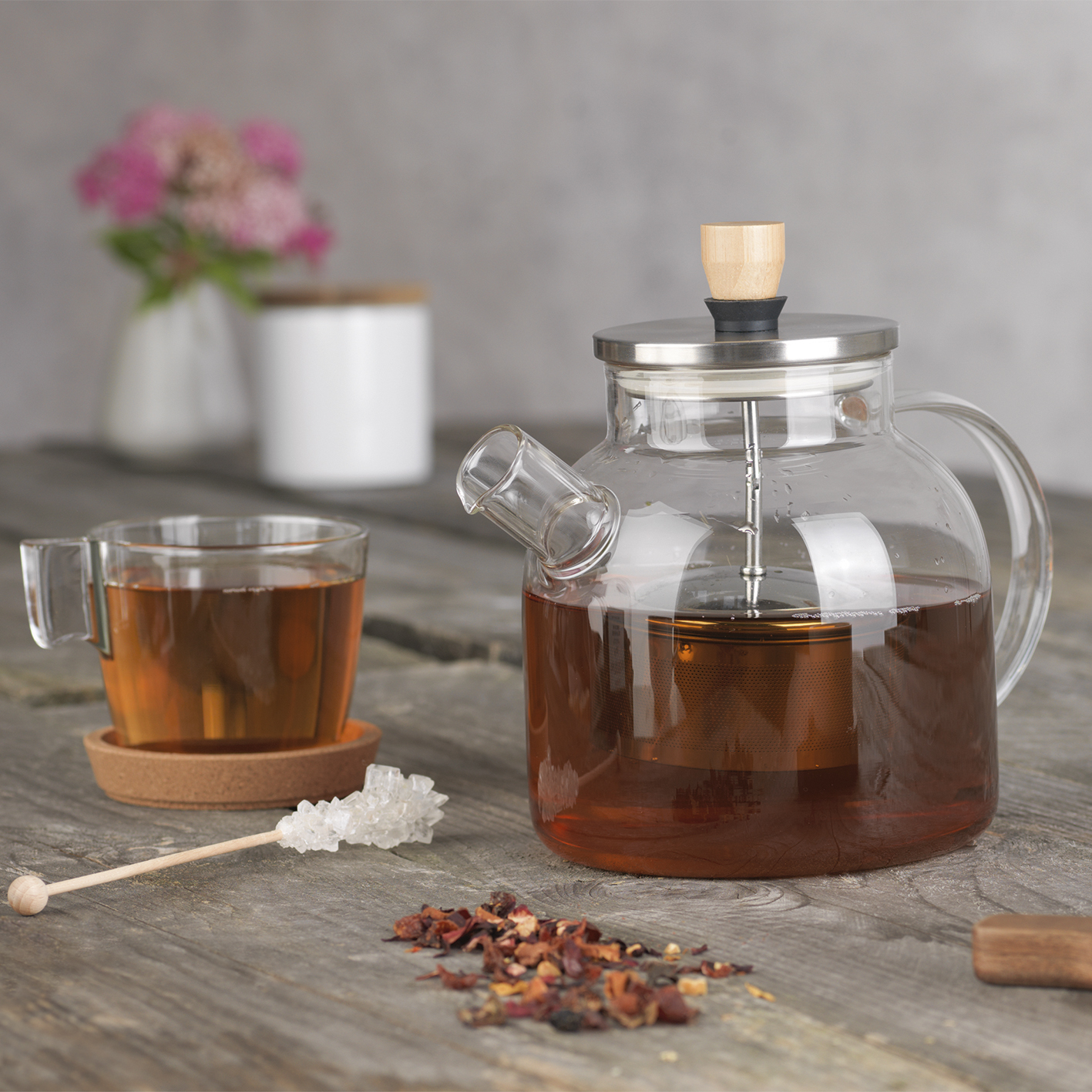 Gläserne Teekanne mit Tee, Tasse, Zucker und Blumen.