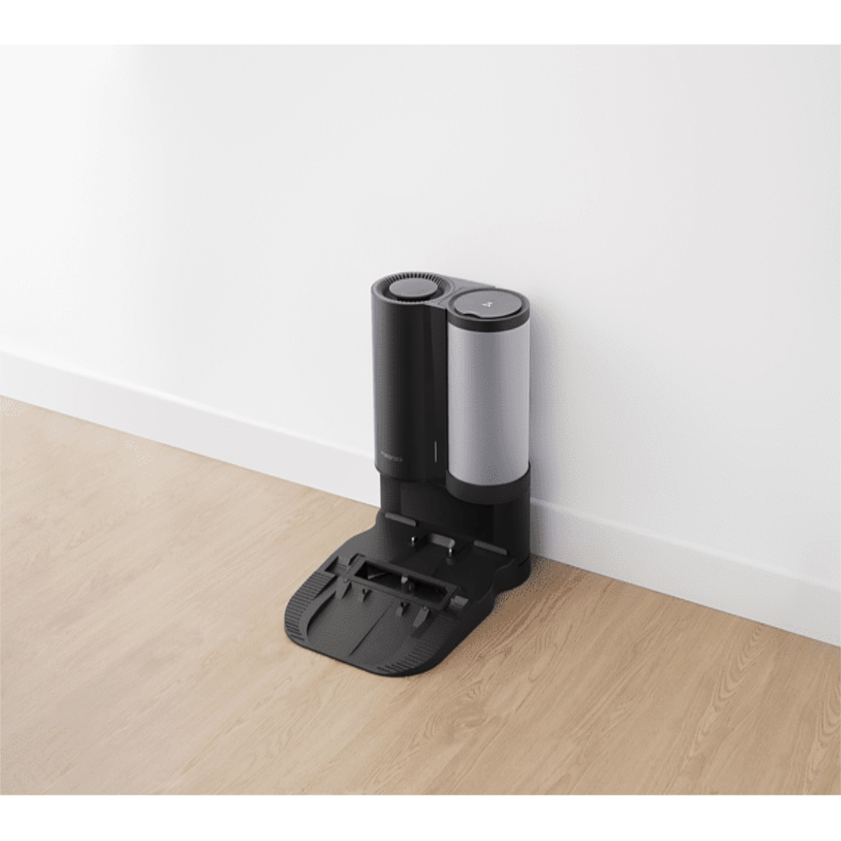 Aspiradora robot negra y gris en piso de madera, contra pared blanca. Se está cargando.