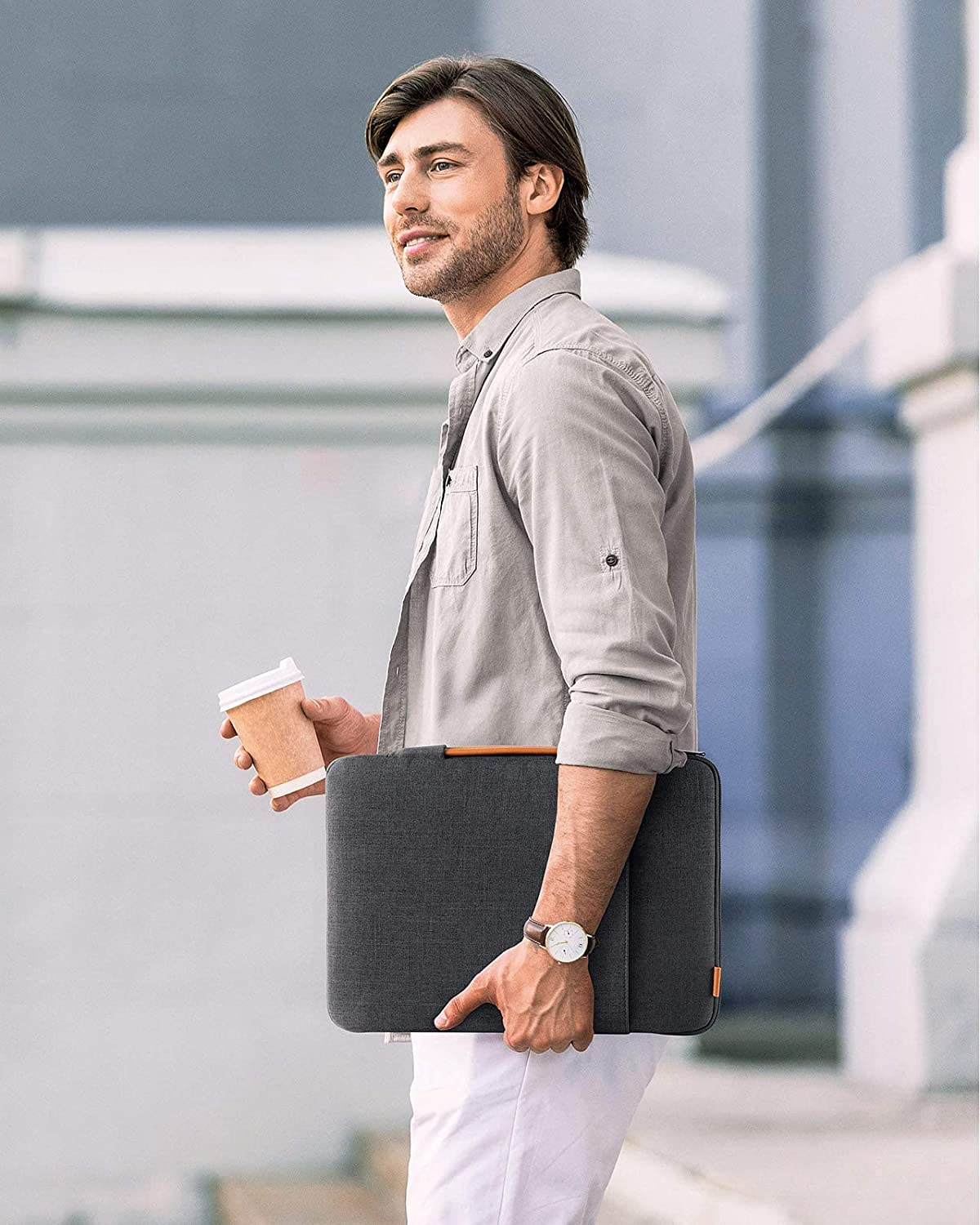 Un hombre con camisa gris y pantalón blanco sostiene una taza de café y una funda para portátil. Sonríe.