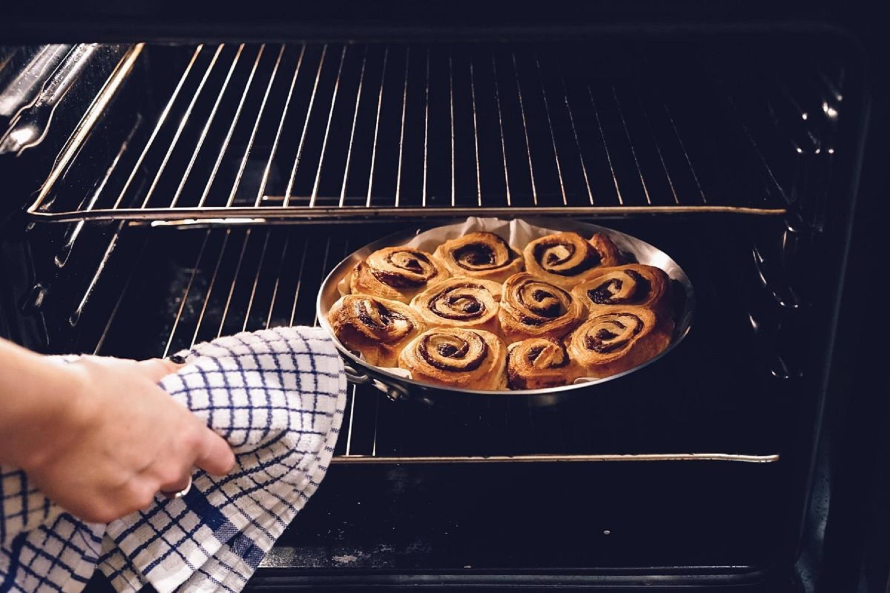 Una bandeja de rollos de canela se está horneando en un horno. Una persona usa una toalla a cuadros para sacarlos.