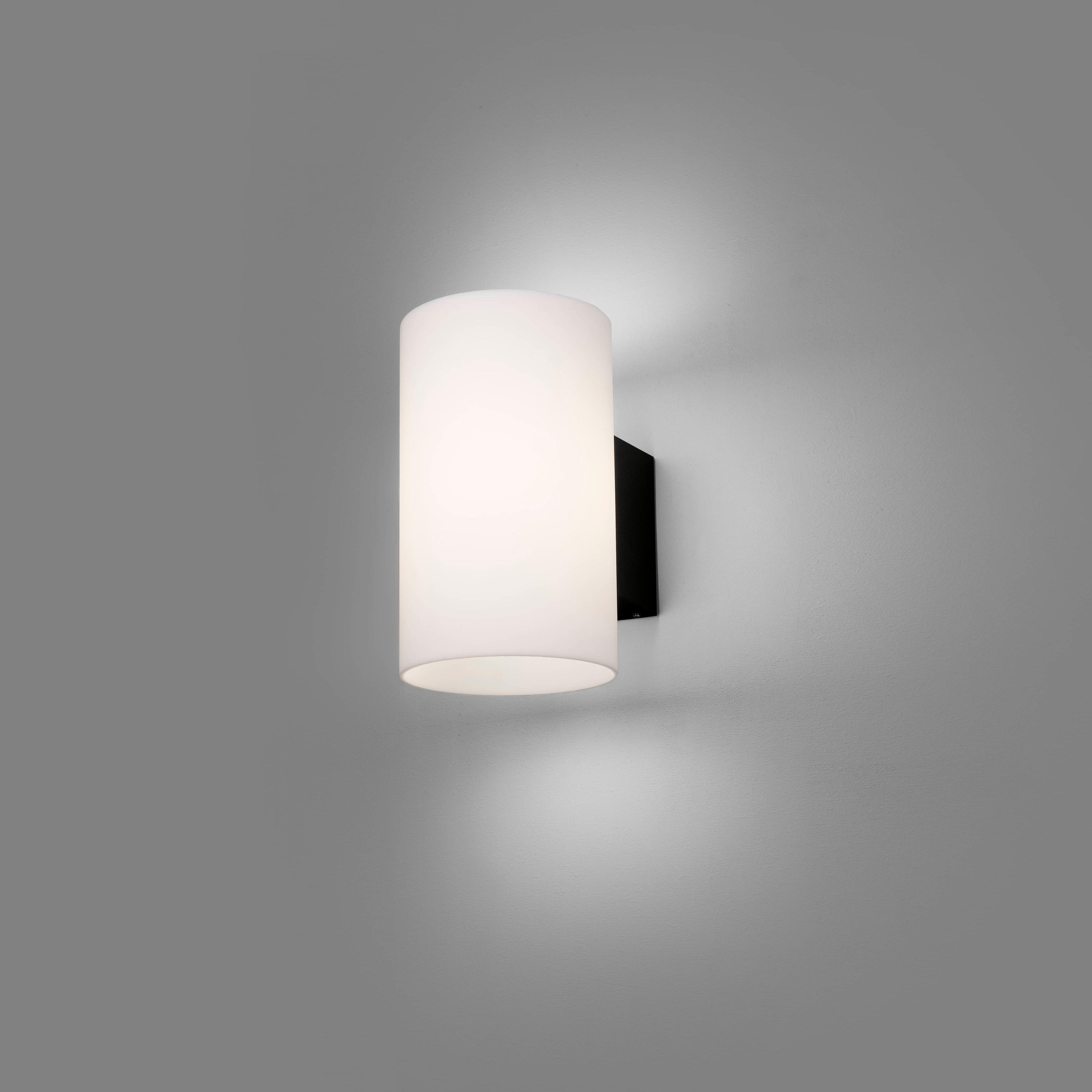 Weißer zylindrischer Wandleuchter an einer grauen Wand. Die Lampe hat eine schwarze Rückseite.