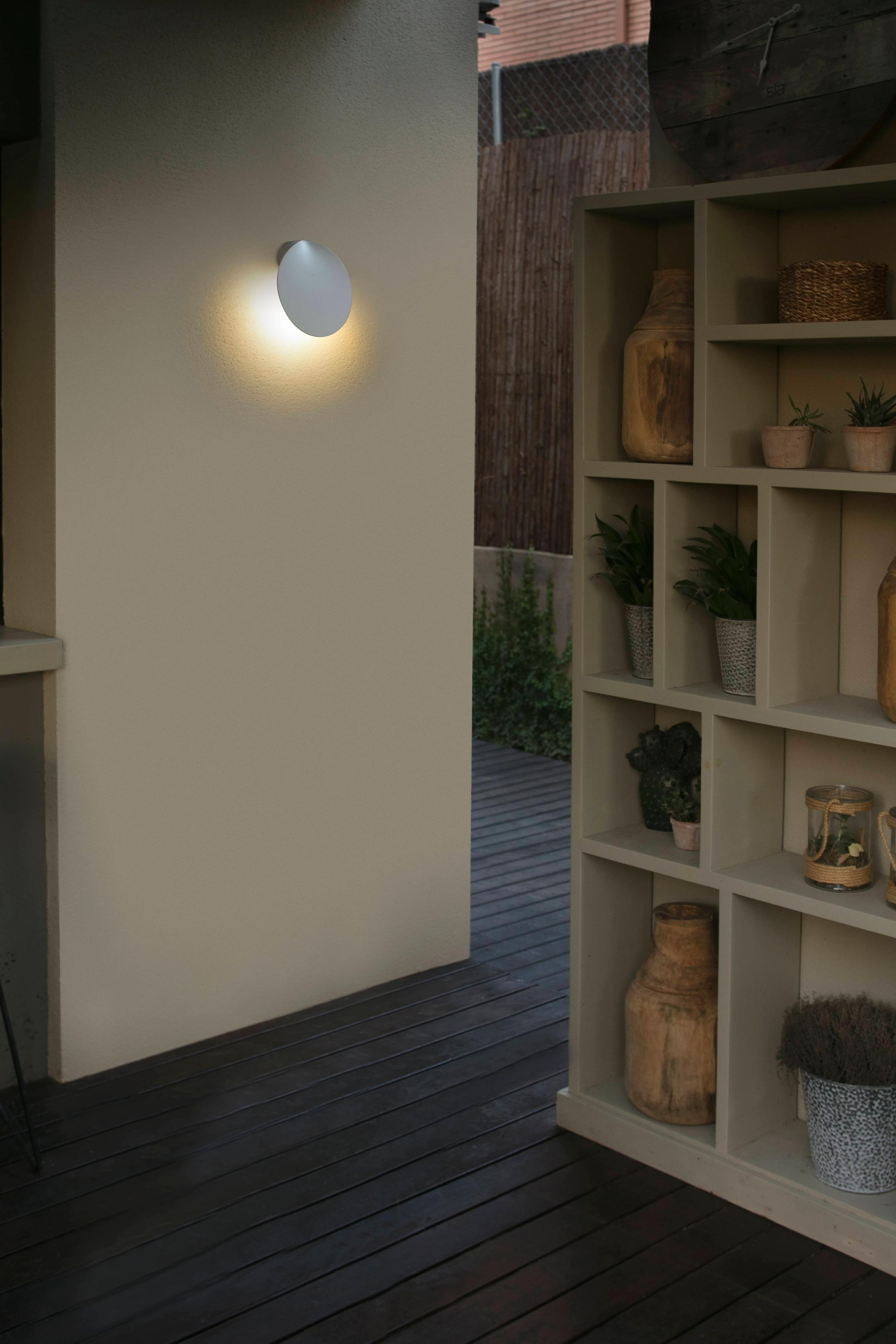 Scena all'aperto con una lampada da parete e una scaffalatura in legno con piante e vasi. La luce illumina il muro.