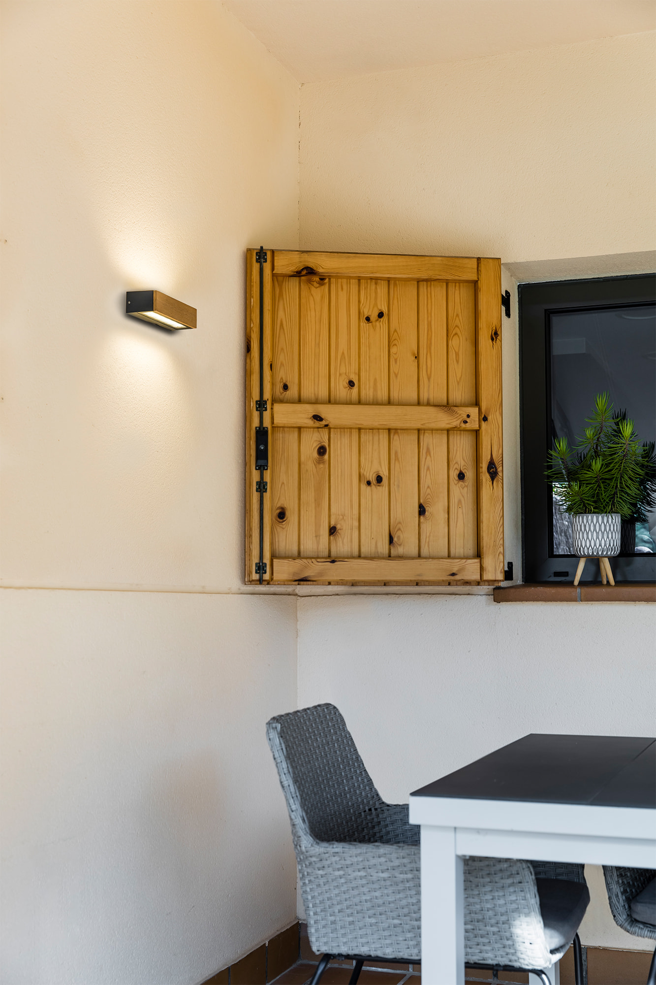 Außenansicht: Holzläden, Außenleuchte, Tisch und Stuhl vor einer beigen Wand.