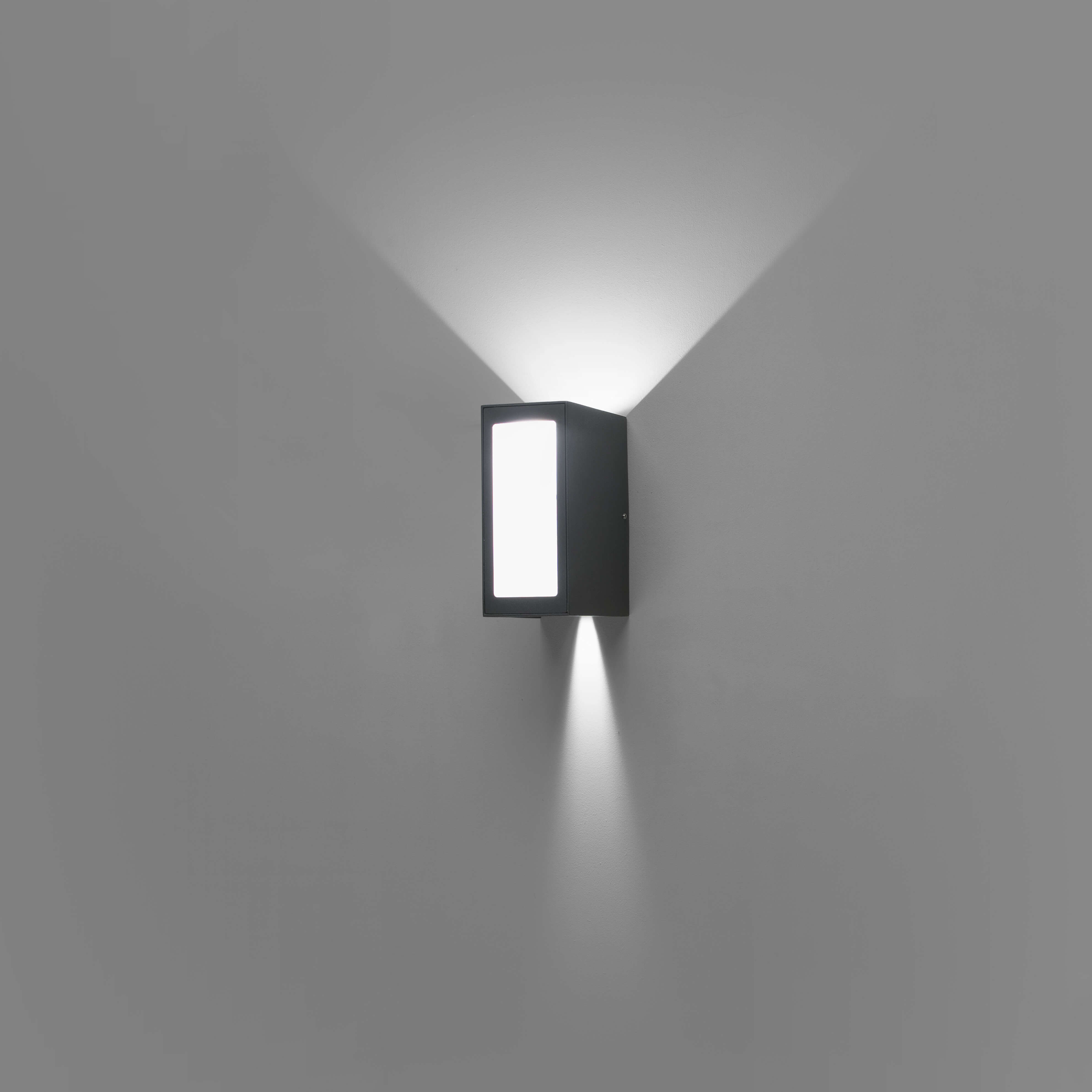 Lampada da parete nera che emette luce verso l'alto e verso il basso su un muro grigio, un rettangolo luminoso al centro.