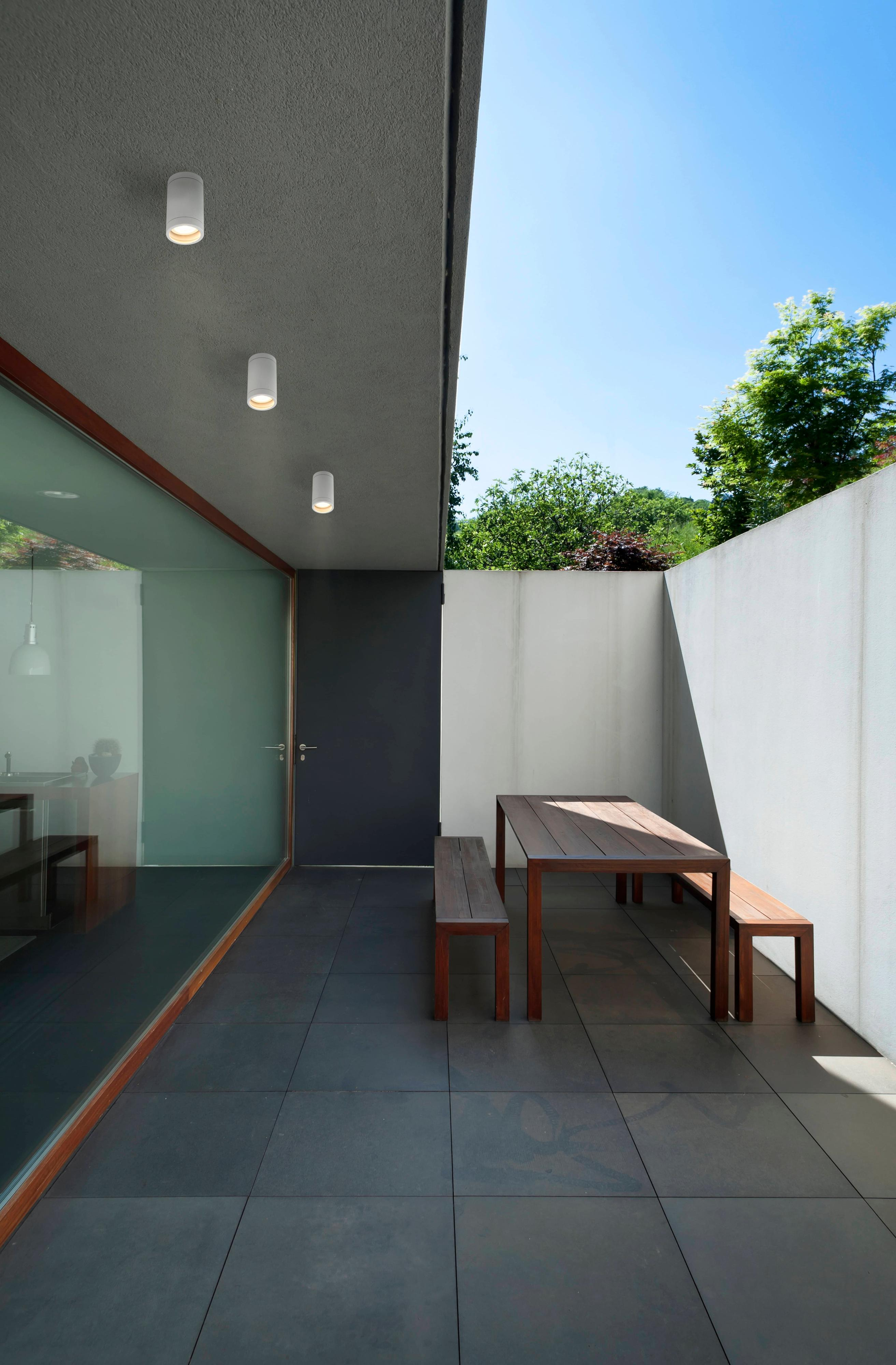 Un entorno exterior moderno con una mesa, bancos y tres luces de techo. Día soleado.