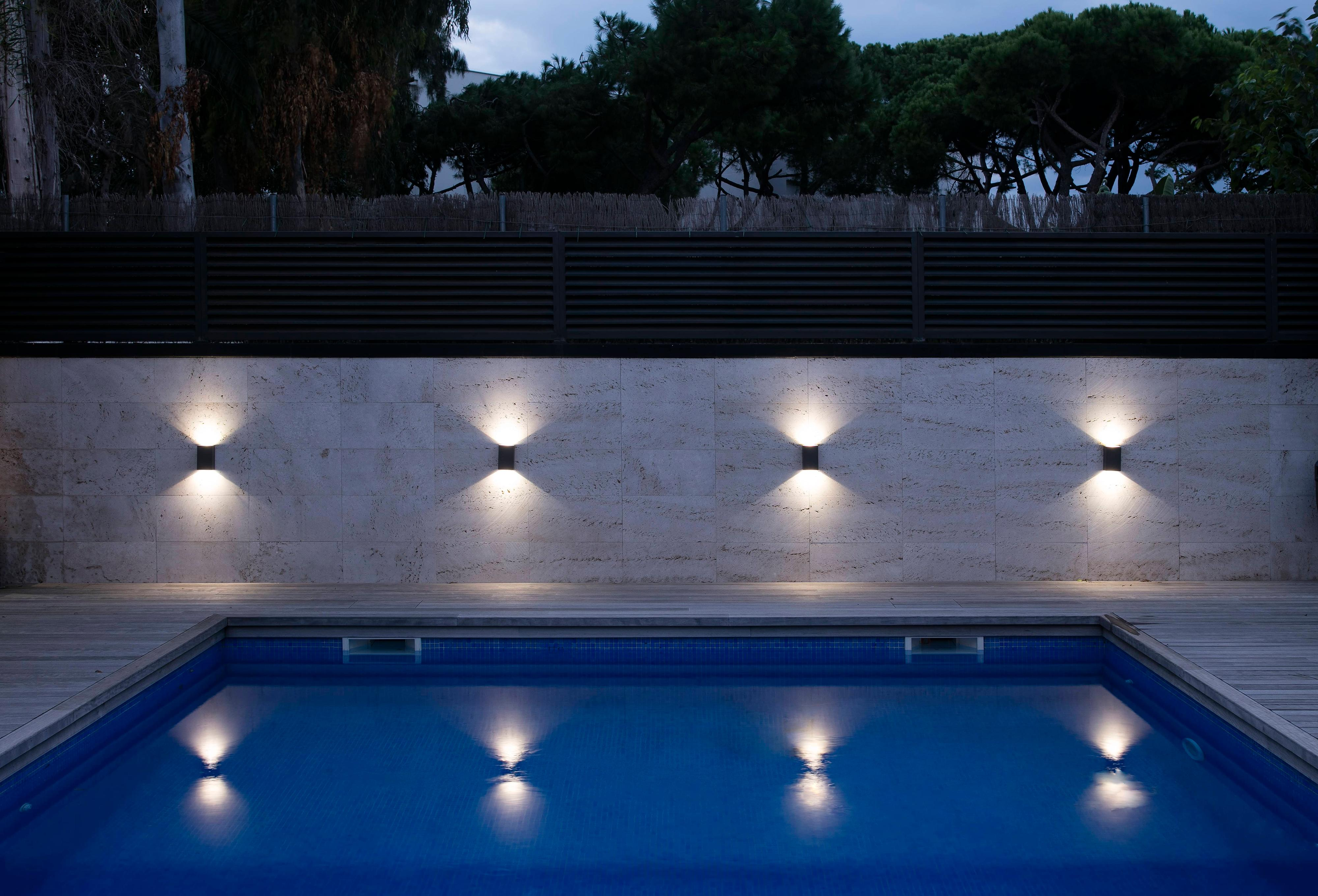 Nachtaufnahme eines Außenpools mit Lichtern. Vier Lichter sind an der Wand, spiegeln sich im Wasser.