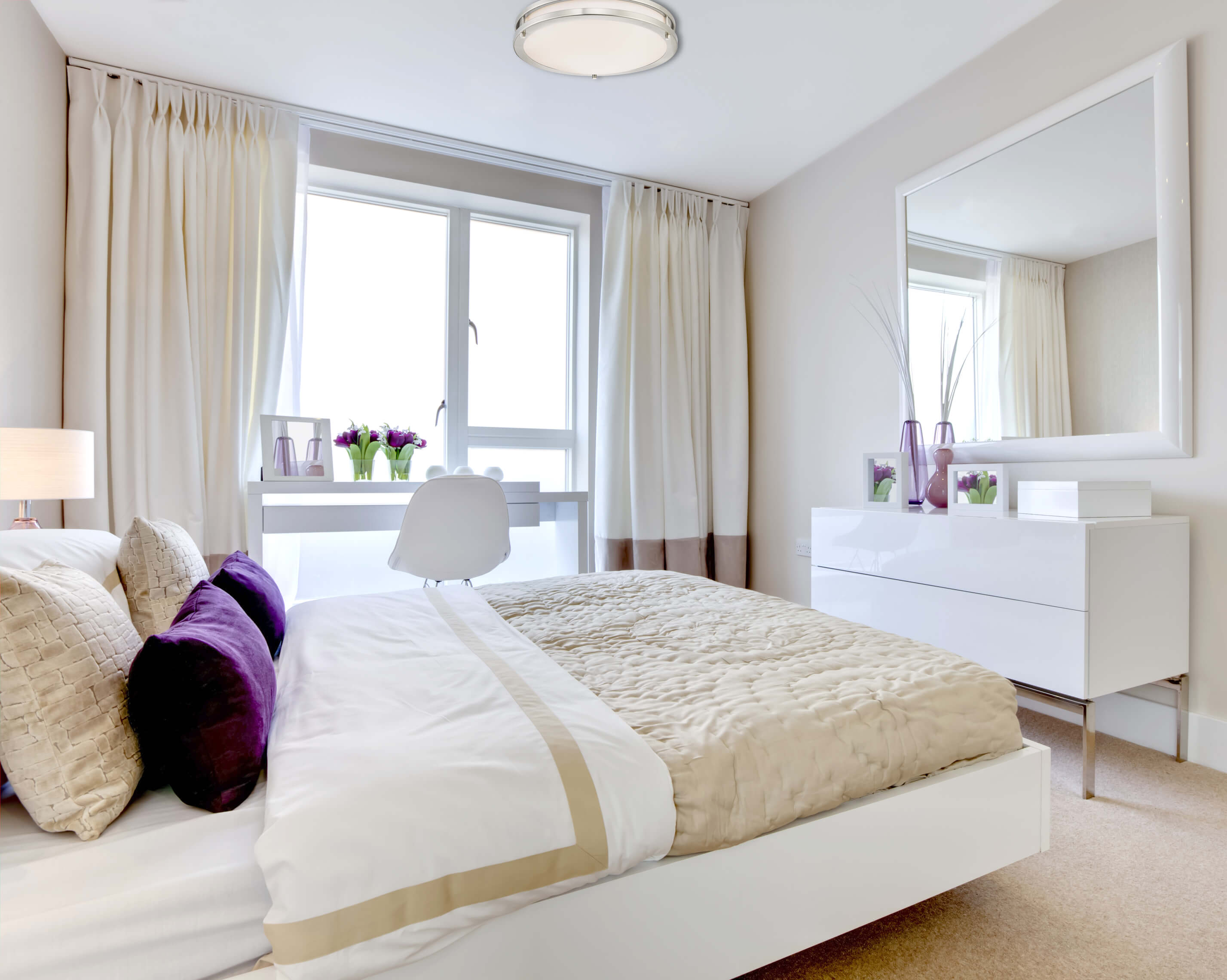Ein Schlafzimmer mit Bett, Kommode, großem Spiegel und Fenster. Das Bett hat Kissen und eine beige Bettdecke. Weiße Vorhänge.