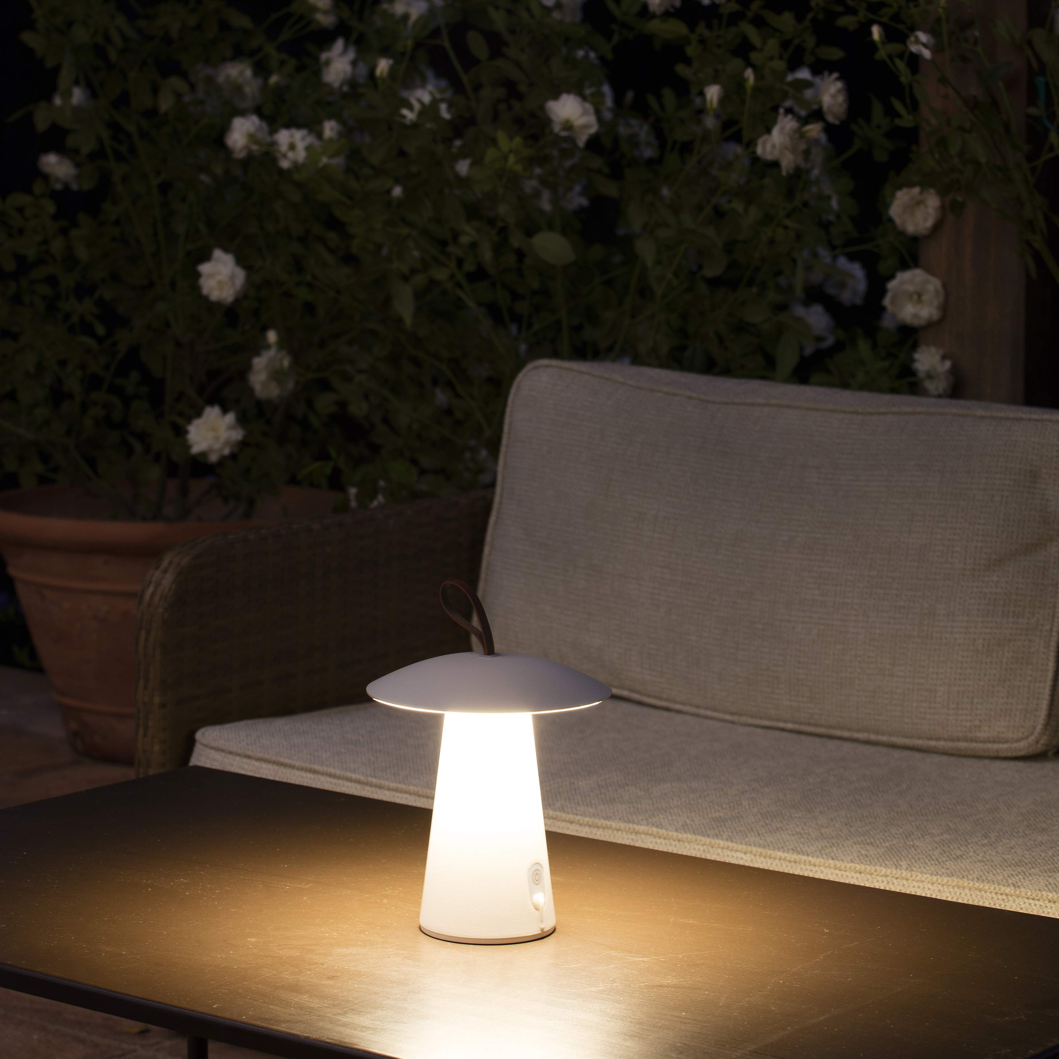 Eine weiße, pilzförmige Außenlampe auf einem Tisch, neben einem Sofa und Pflanzen bei Nacht.