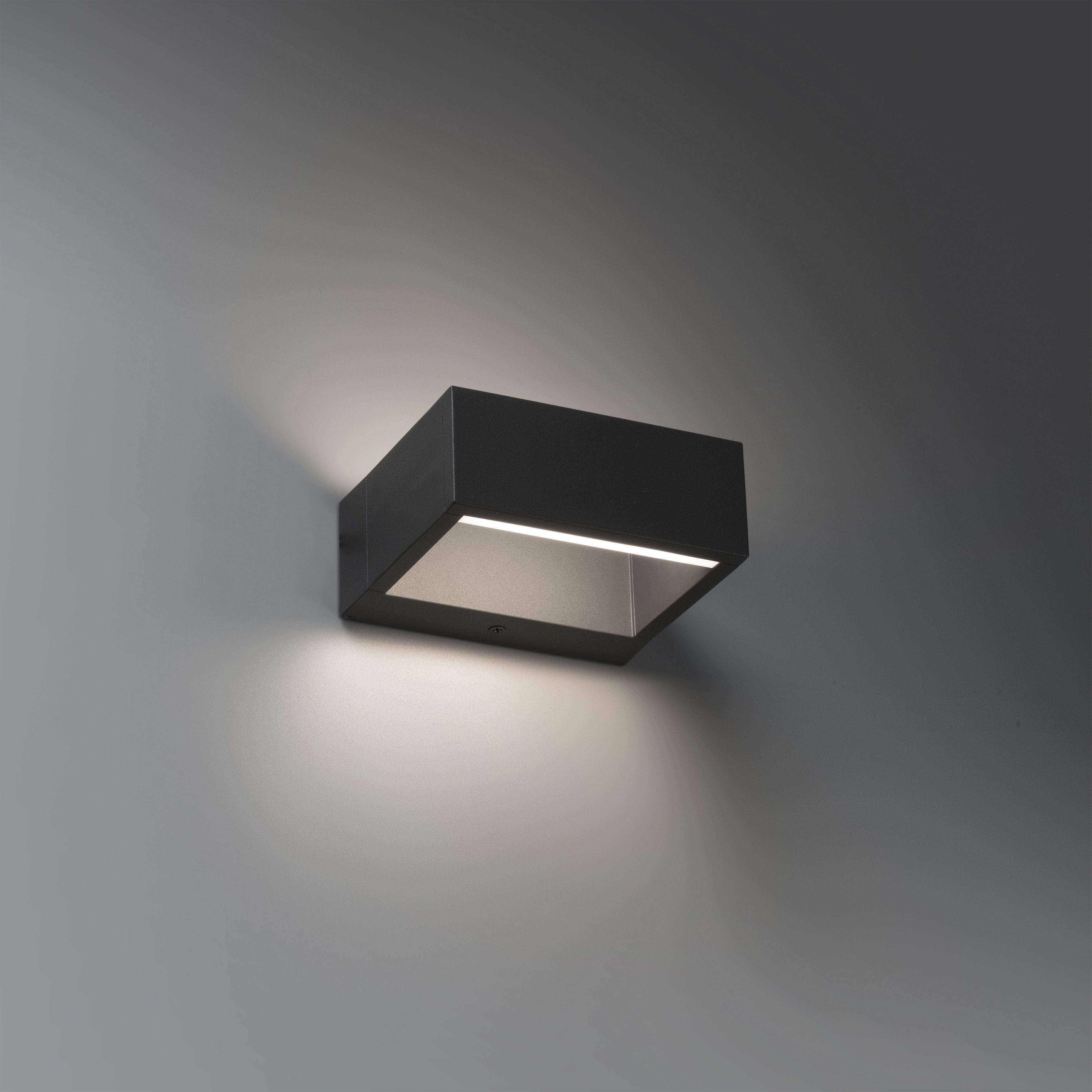Eine schwarze, rechteckige Wandleuchte ist an einer grauen Wand befestigt. Das Licht scheint nach unten.