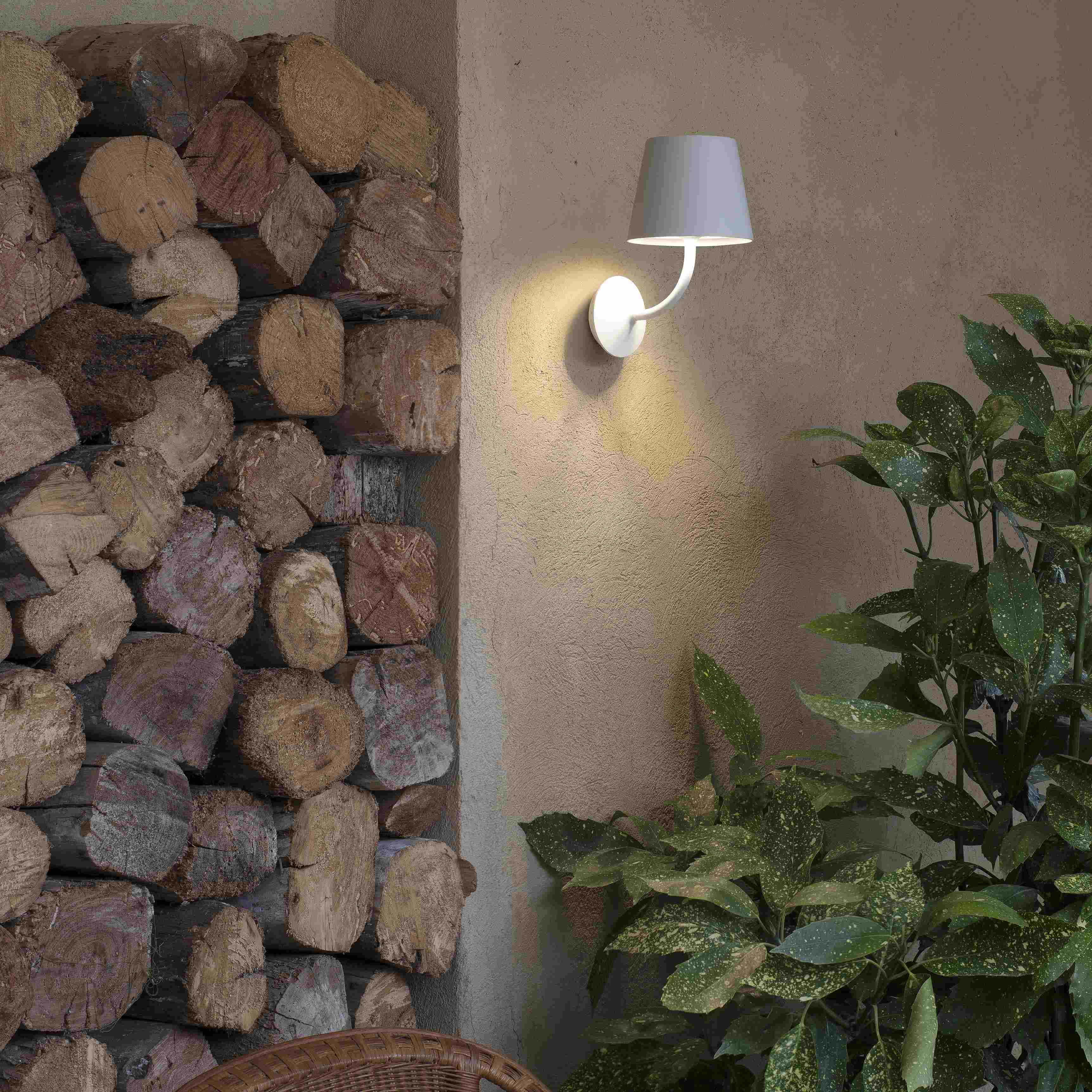 Lampada da parete per esterni con paralume e una catasta di legna sullo sfondo. Sono visibili piante verdi.