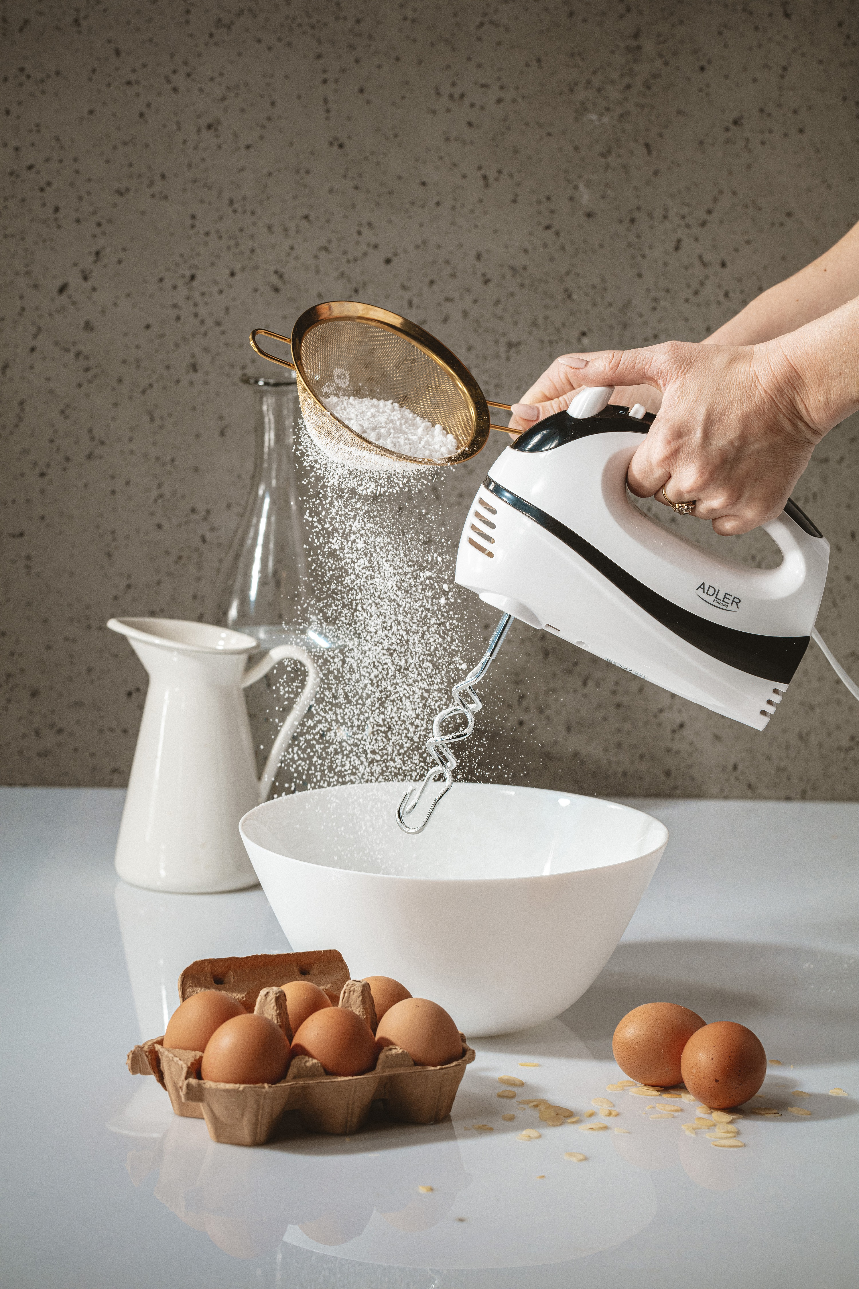Osoba przesiewająca cukier puder do miski z jajkami i mikserem.