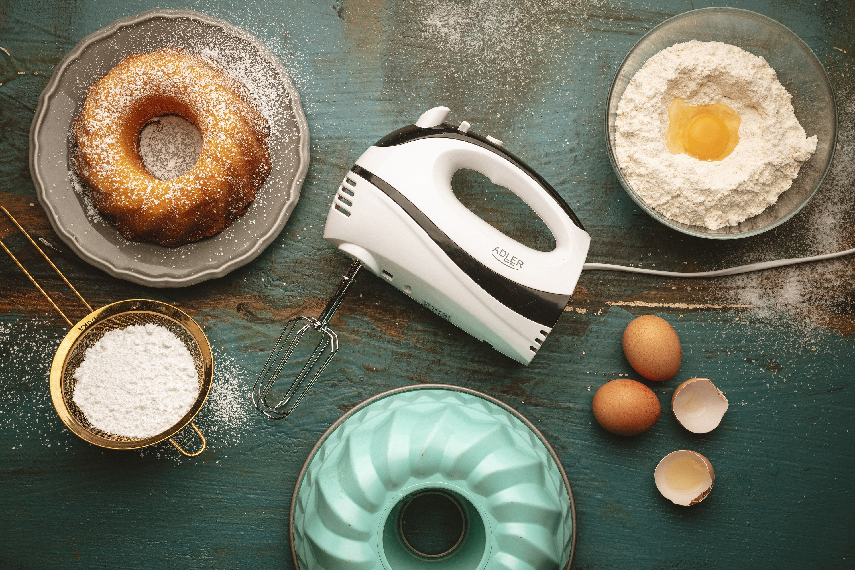 Widok z góry na akcesoria do pieczenia. Zawiera ciasto, mąkę, jajka i mikser ręczny na niebiesko-zielonej powierzchni drewnianej.