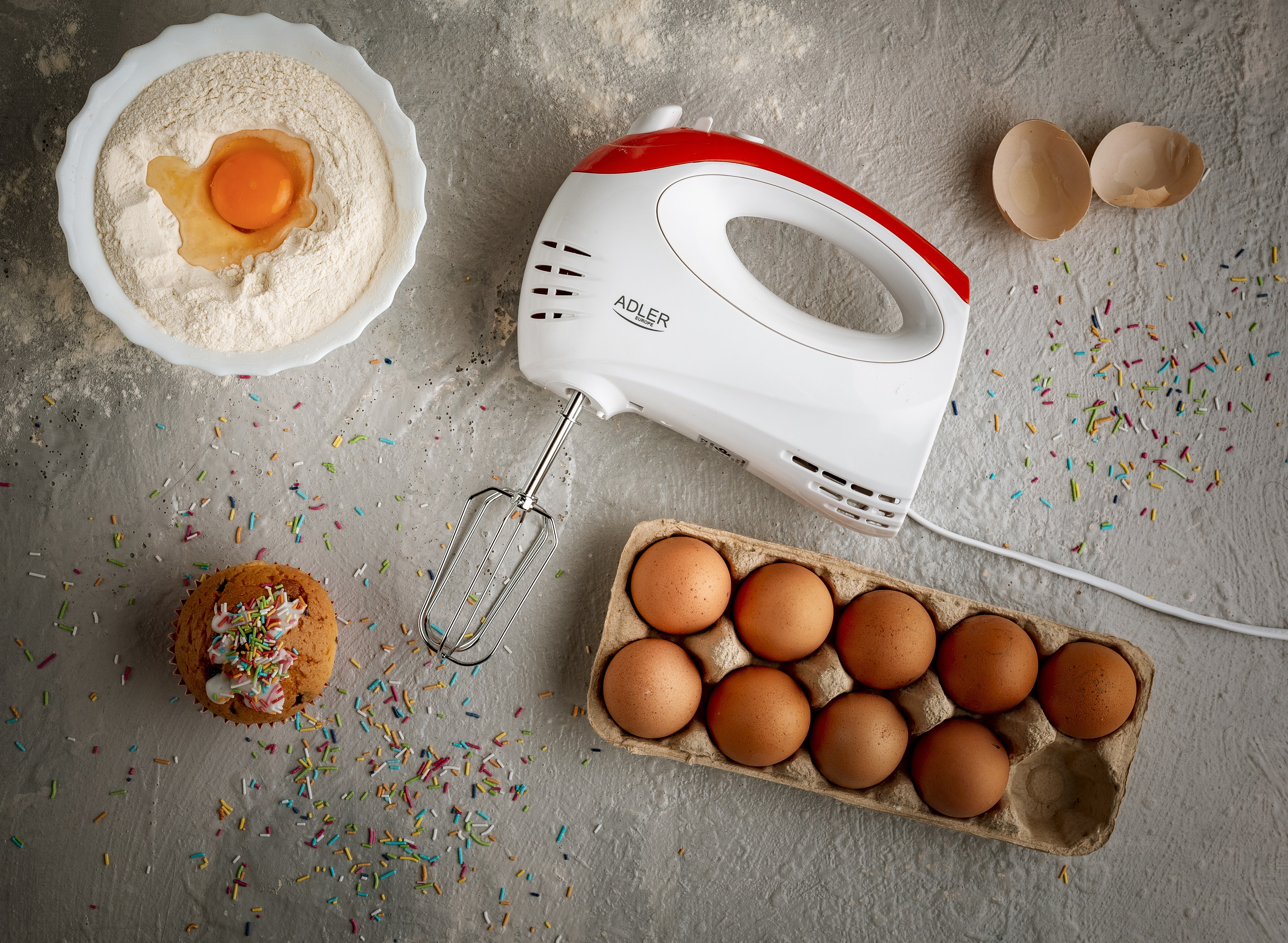 Scena mieszania: miska z mąką i jajkiem, mikser, karton jajek i babeczka z posypką.