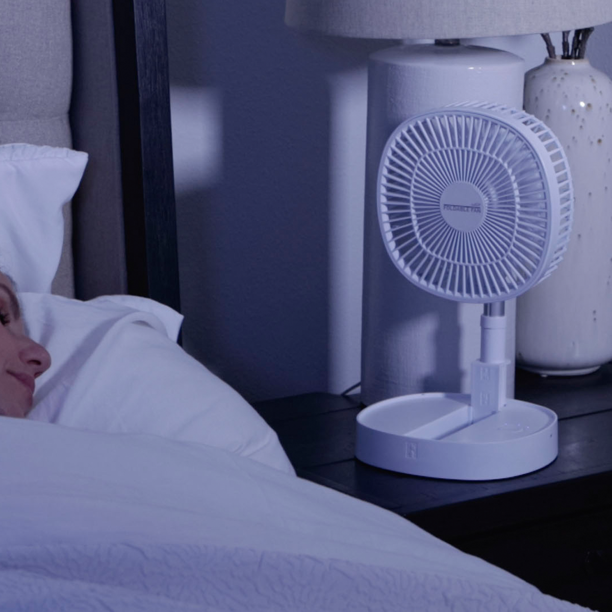 Weißer Klappventilator auf einem Nachttisch neben einem Bett. Eine Frau schläft unter einer weißen Decke.
