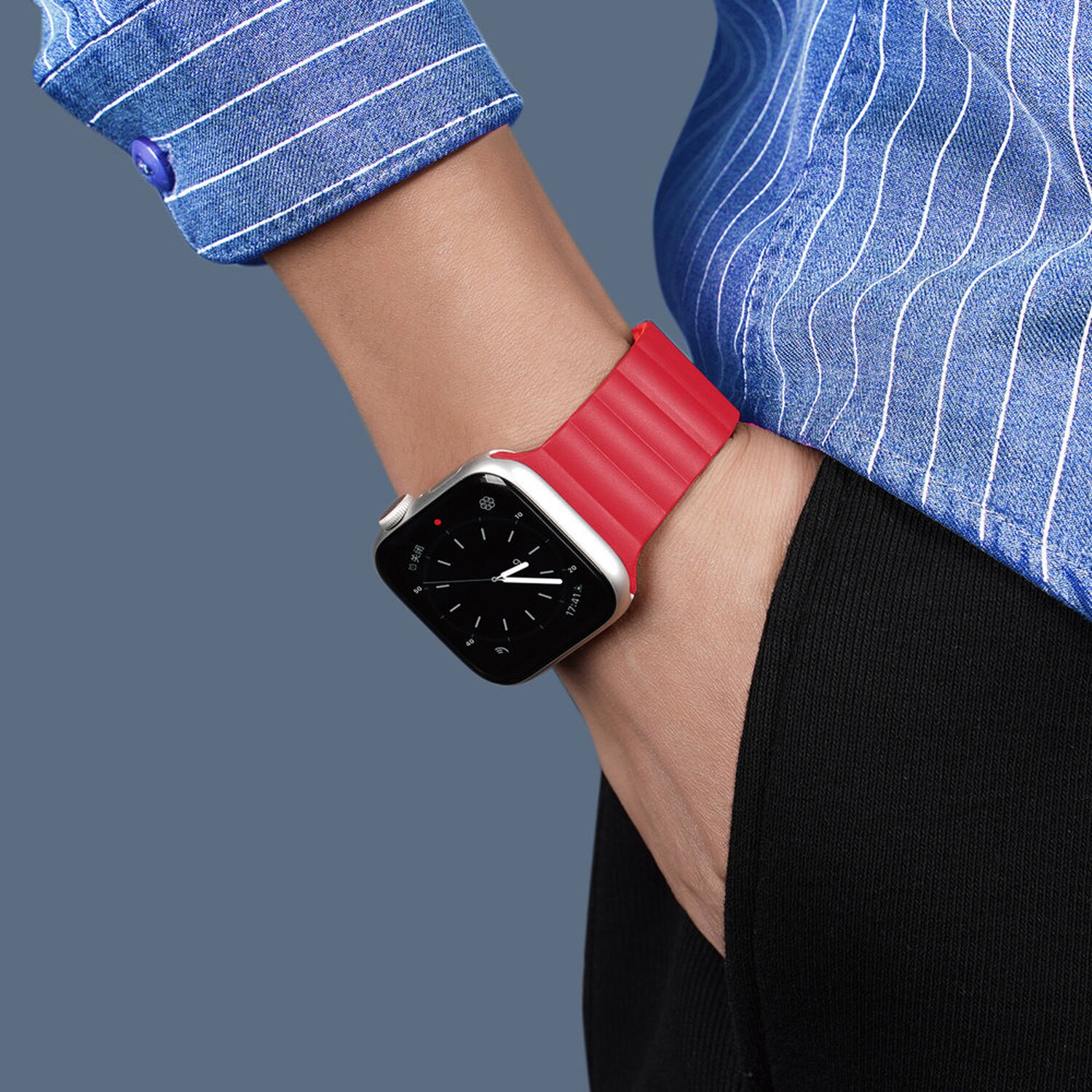 Handgelenk mit silberner Apple Watch und rotem Armband vor blauem Hintergrund.