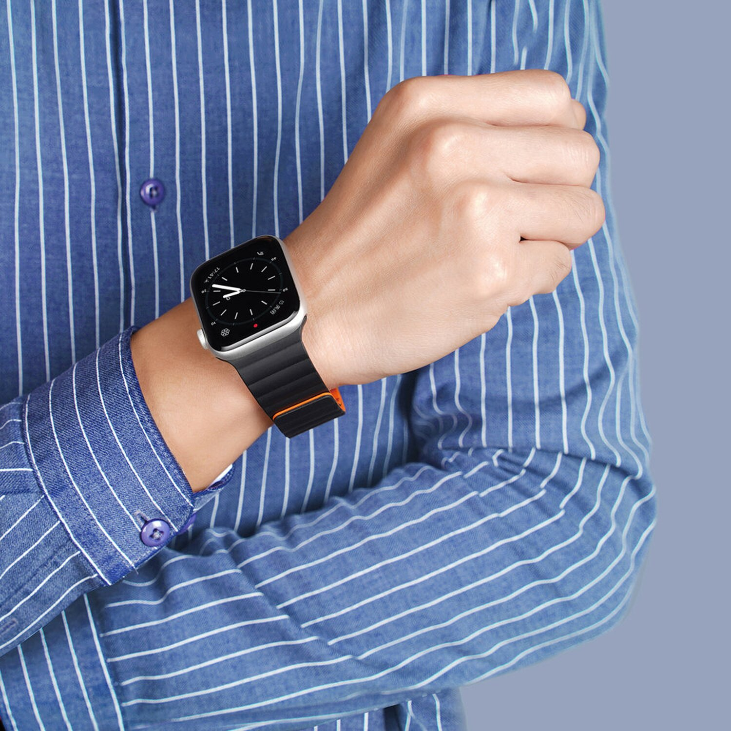 Das Handgelenk einer Person mit einer Smartwatch mit schwarzem und orangenem Armband, vor einem blau-weiß gestreiften Hemd.