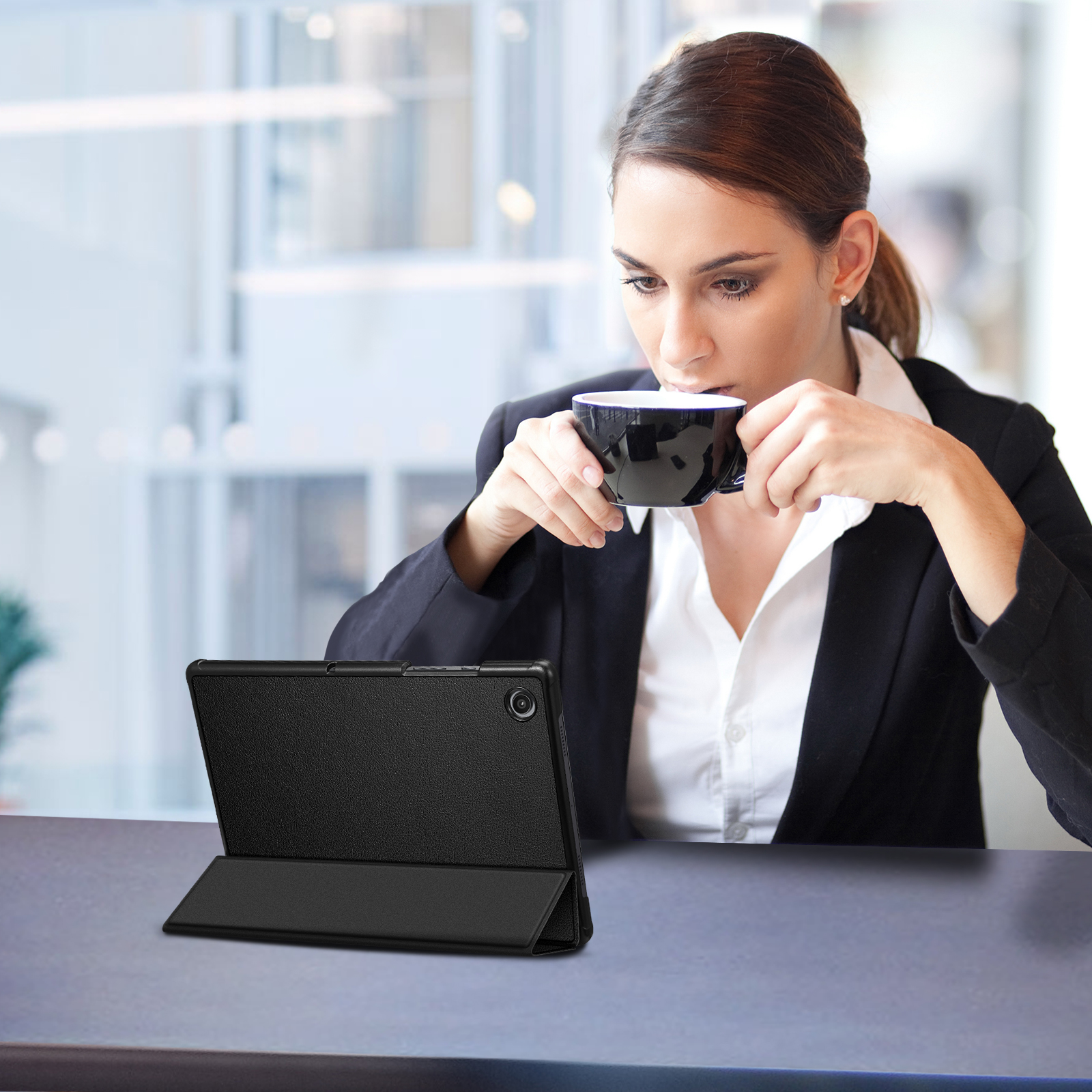 Eine Frau trinkt aus einer schwarzen Tasse, während sie an einem Tisch sitzt. Ein Tablet mit Hülle liegt auf dem Tisch.