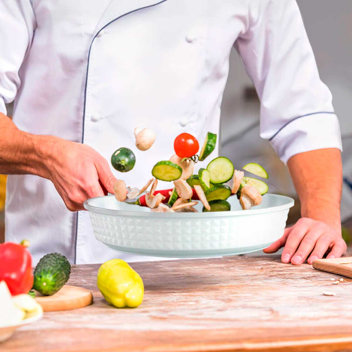 Un chef lanzando verduras en un cuenco blanco. Se ven varias verduras frescas.