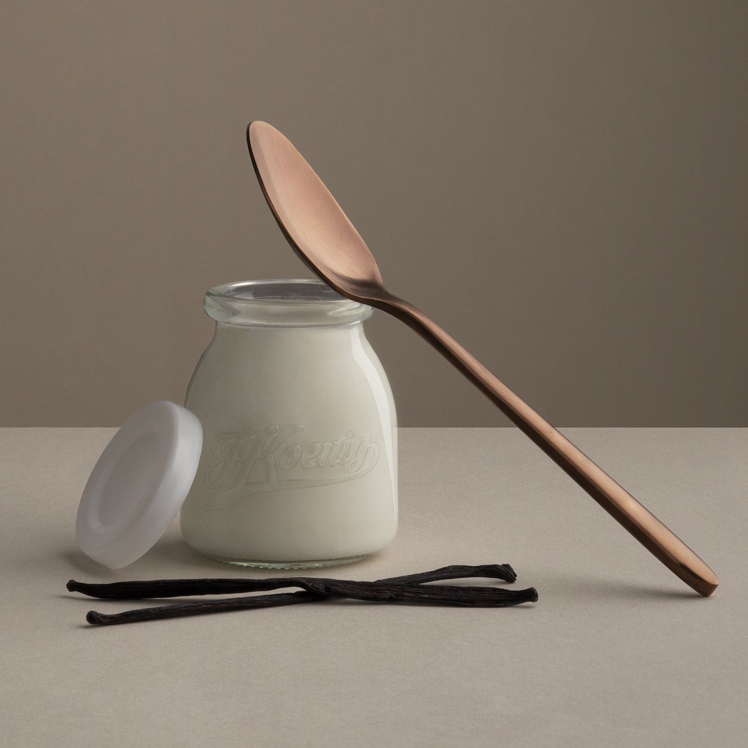 Szklany słoik z mlekiem, łyżką i laskami wanilii. Tło w neutralnym beżowym kolorze.