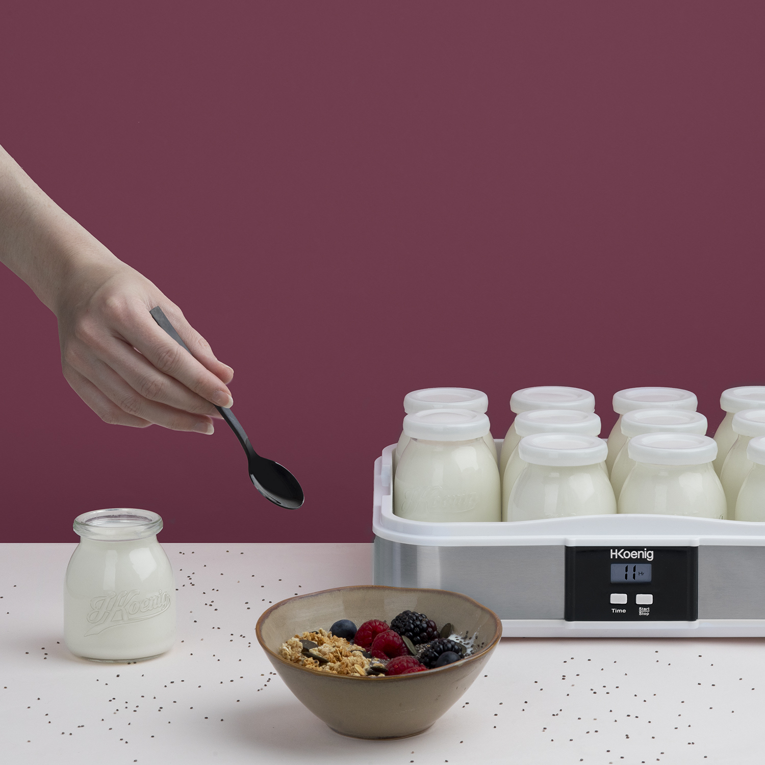 Una mano che tiene un cucchiaio sopra vasetti di yogurt, granola e bacche su un tavolo, con una yogurtiera.