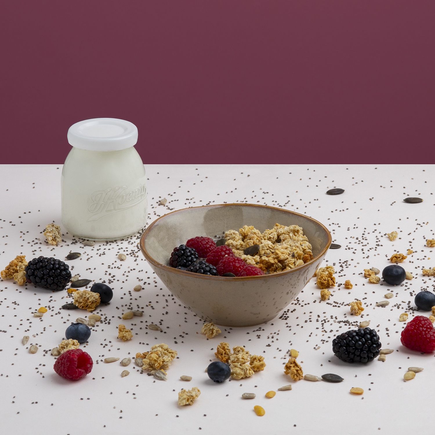 Ciotola di cereali con frutti di bosco, bottiglia di latte e semi sparsi. Sfondo viola, superficie bianca.