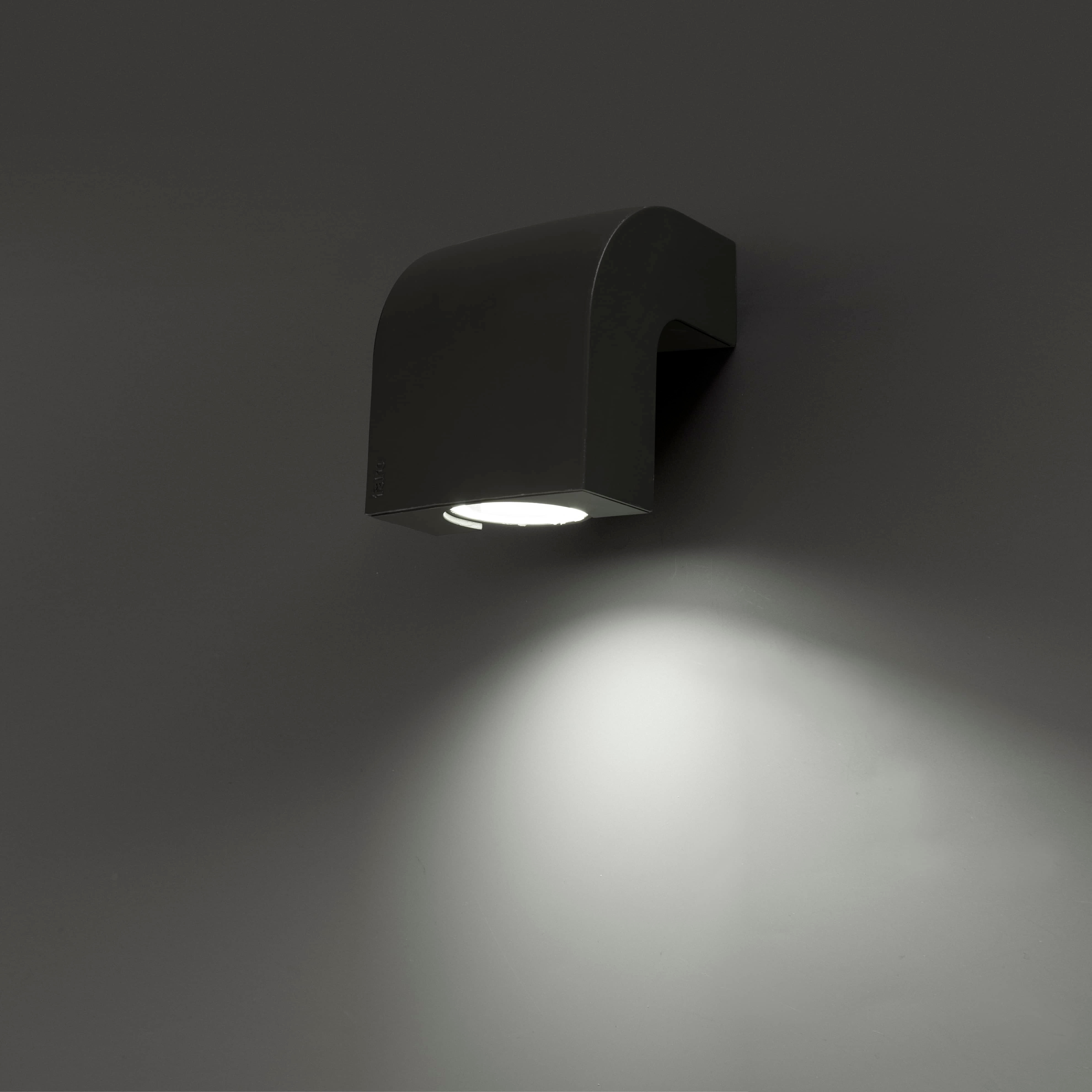 Eine schwarze, wandmontierte Außenleuchte. Sie wirft einen nach unten gerichteten Lichtkegel auf die graue Wand.