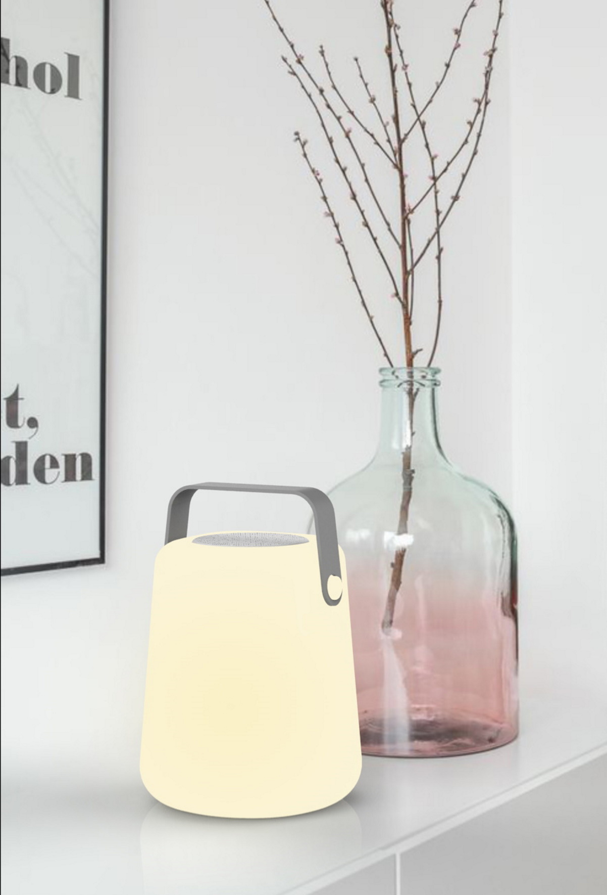 Eine leuchtende tragbare Lampe mit Griff steht auf weißer Oberfläche. Eine Vase mit Zweigen dahinter.