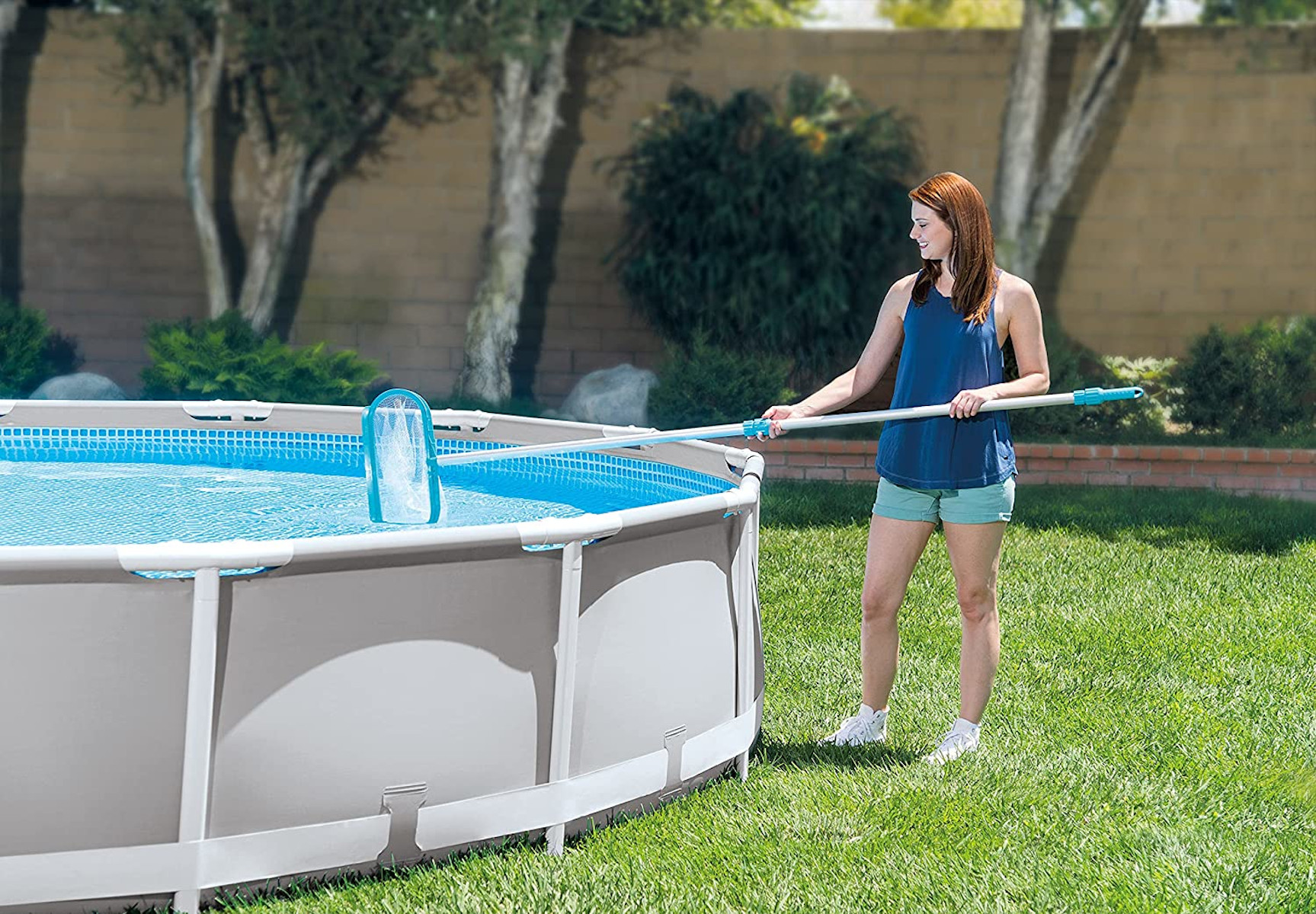 Eine Frau reinigt einen Pool mit einem Netz, steht auf Gras, mit einem Pool im Hintergrund.