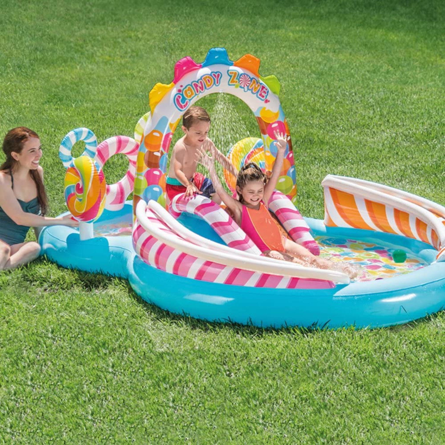 Bambini che giocano in una piscina a tema caramelle con scivolo, acqua e una donna che guarda.