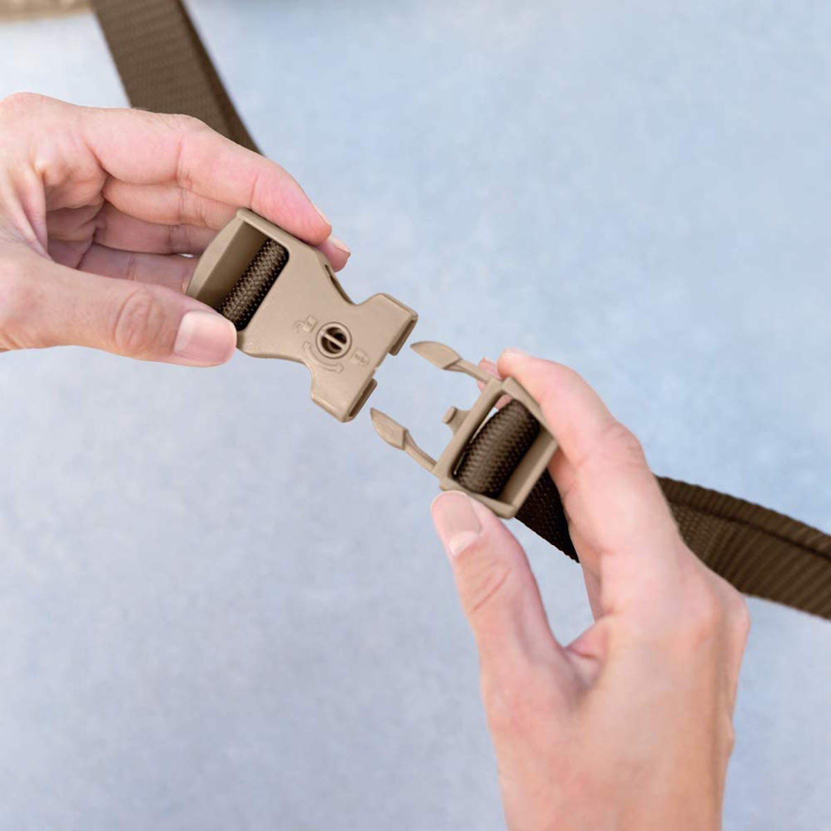 Dłonie łączące beżową klamrę z brązowym paskiem. Tło to jasna, lekko teksturowana powierzchnia.