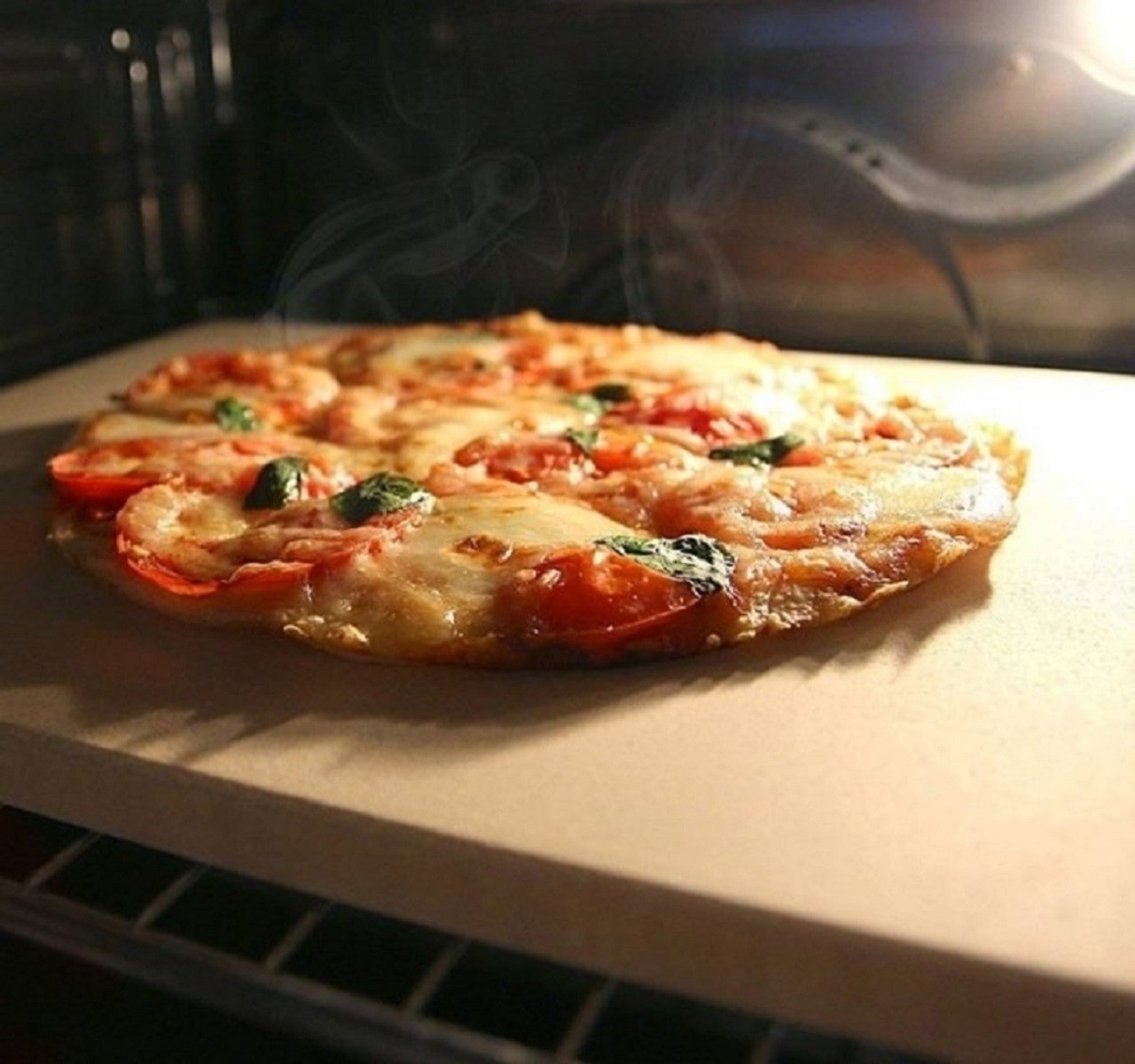 Pizza cocinándose en un horno. Tiene queso, tomates y albahaca. El horno está iluminado, en piedra.