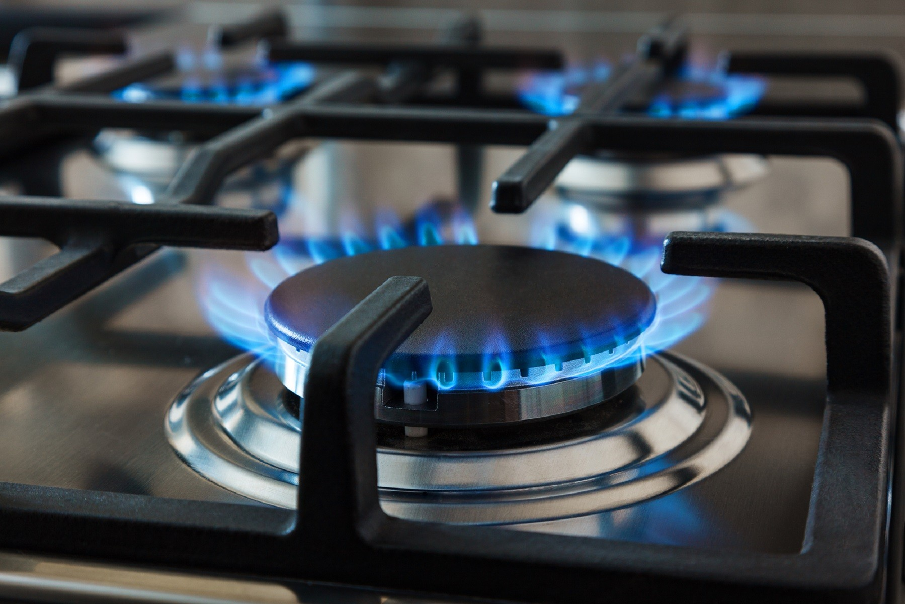 Una estufa de gas con cuatro quemadores. Se ven llamas azules en los quemadores. La estufa es de acero inoxidable.