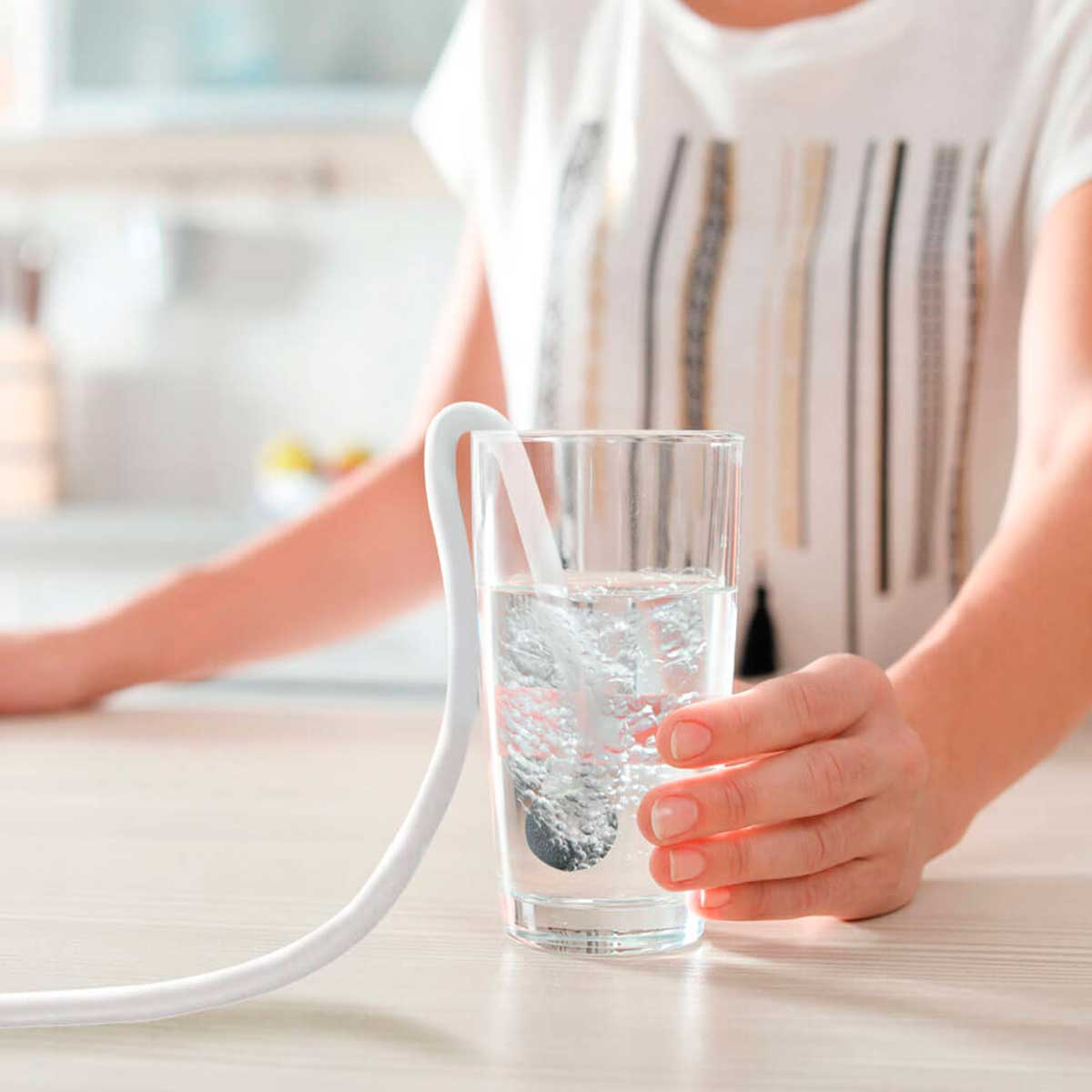 Una persona sostiene un vaso con agua y un tubo blanco. Burbujas suben en el agua. Una cocina está en el fondo.