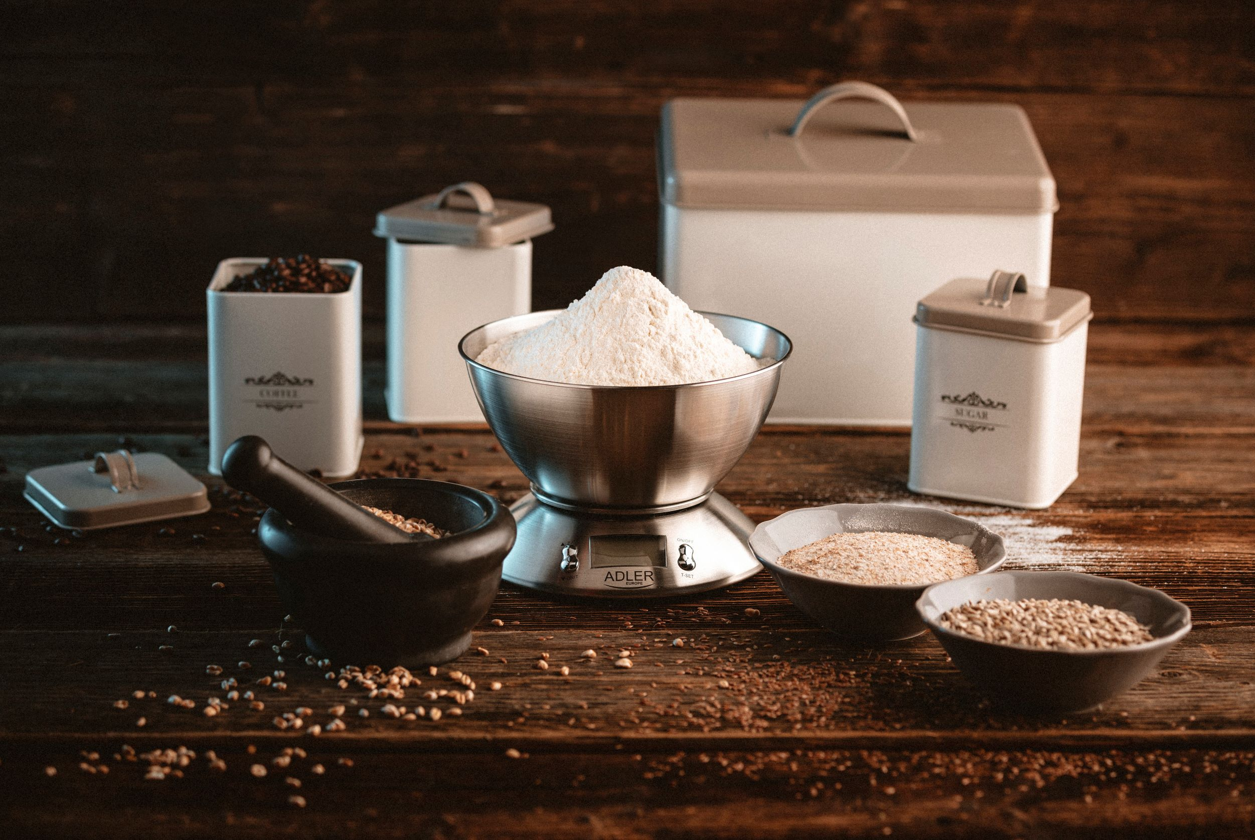 Waga z mąką, otoczona pojemnikami z kawą, cukrem i nasionami, na drewnianej powierzchni.