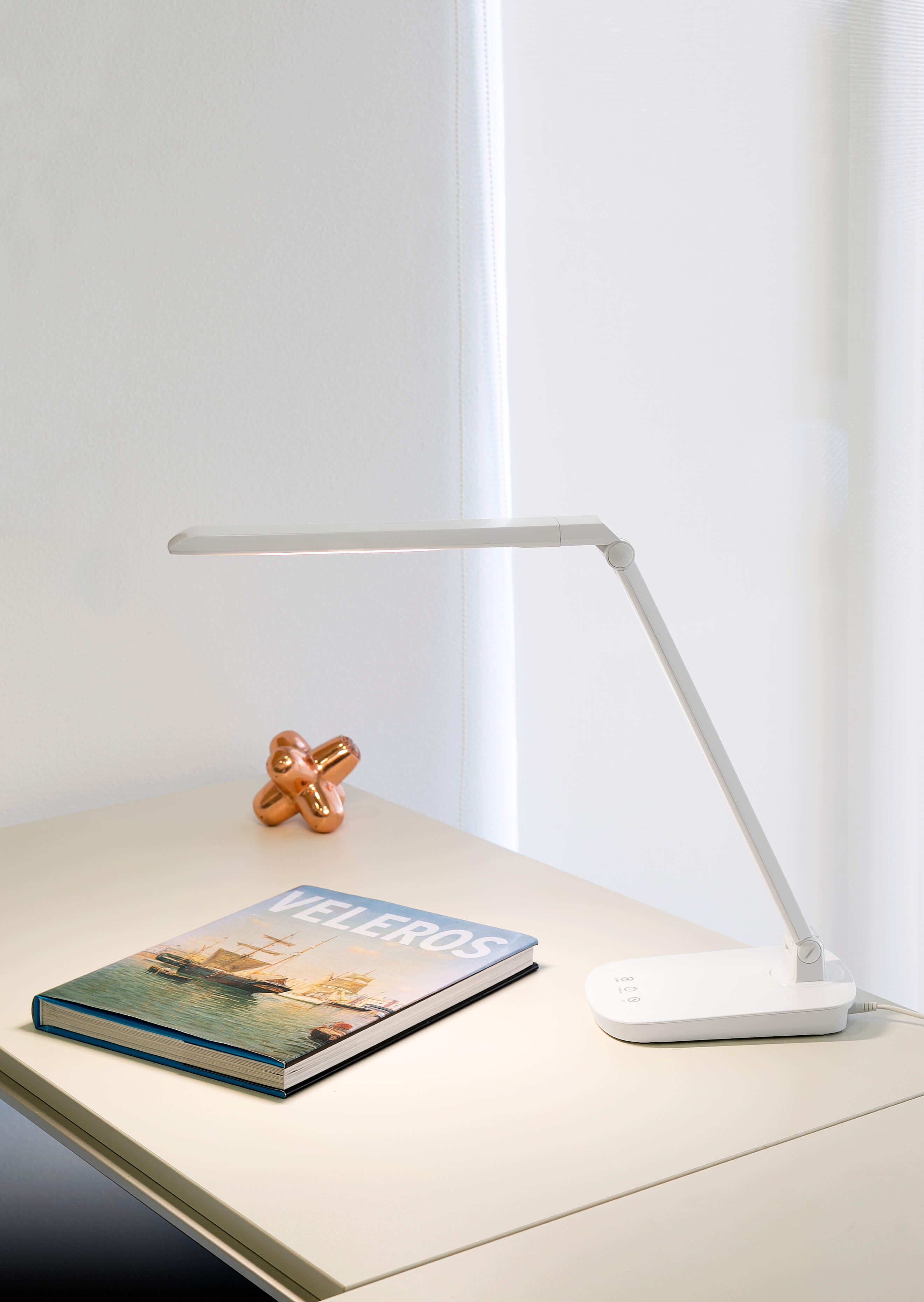 Lampada da scrivania bianca su una scrivania con un libro e una scultura in rame. Sfondo bianco.