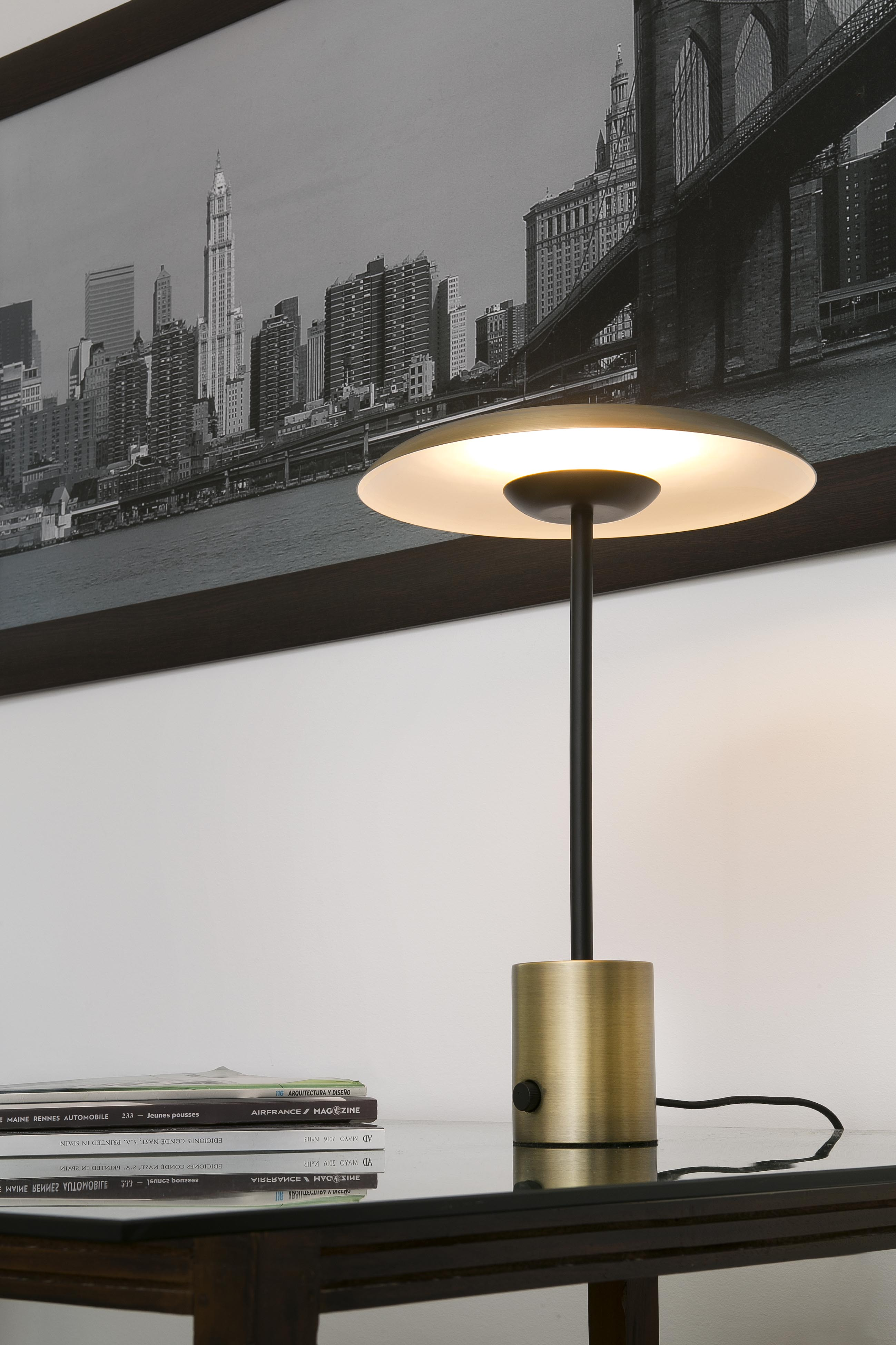 Una lampada con base dorata e paralume circolare su un tavolo di vetro. Sfondo: paesaggio urbano.
