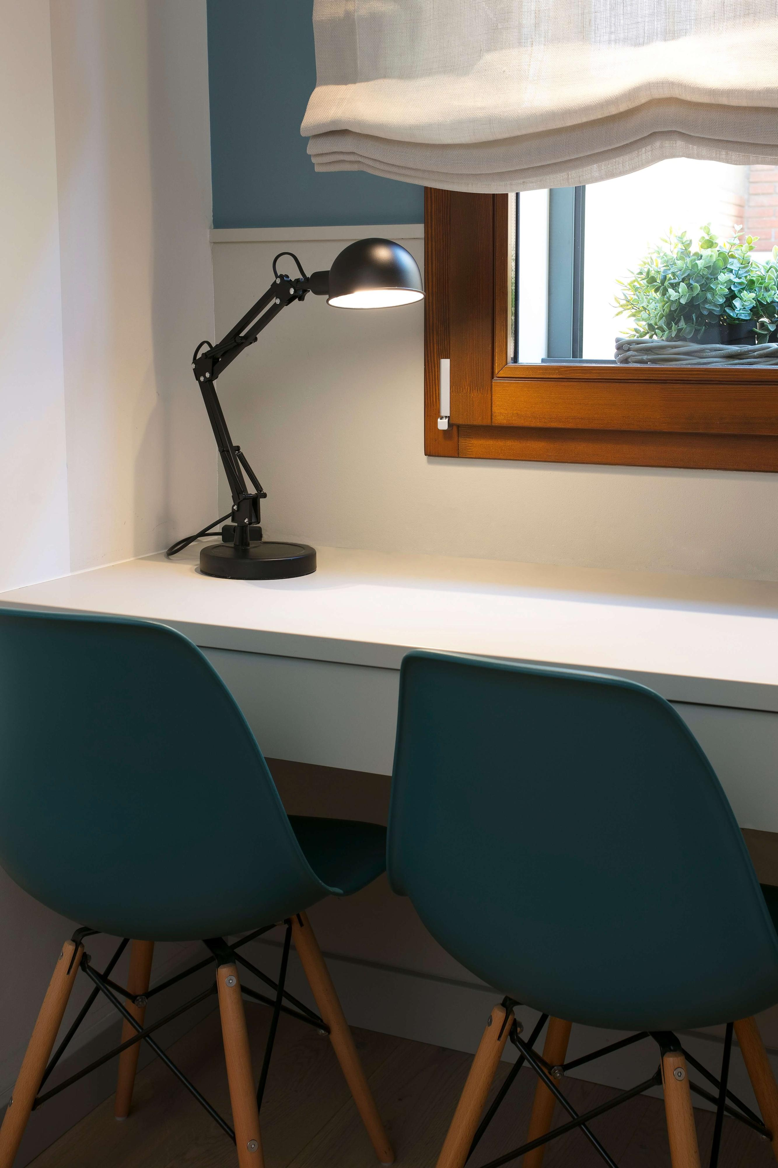 Schreibtisch mit zwei grünen Stühlen, einer schwarzen Lampe, einem Fenster mit Pflanze. Innenszene.